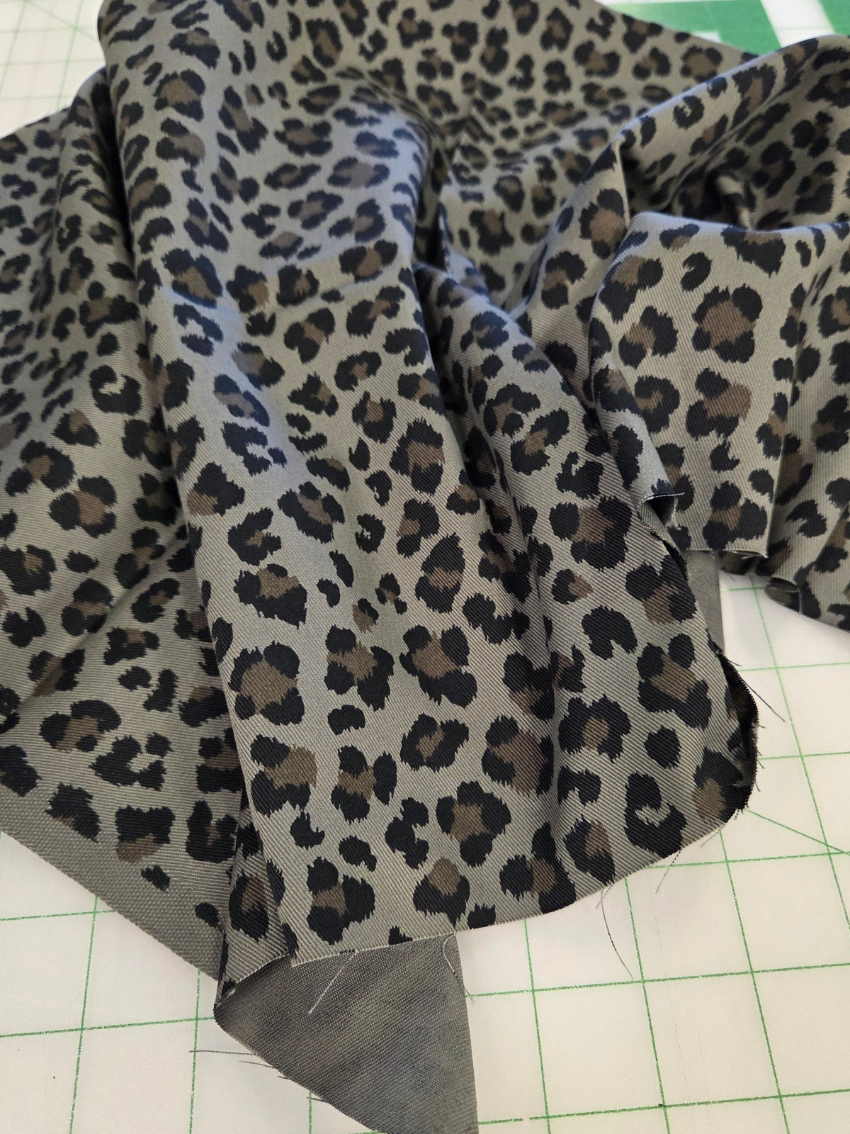 A close-up of leopard-print fabric draped on a white cutting mat with green grid lines. The fabric has a gray base with black and dark brown leopard spots, and a solid gray fabric piece is visible at the folded edge.