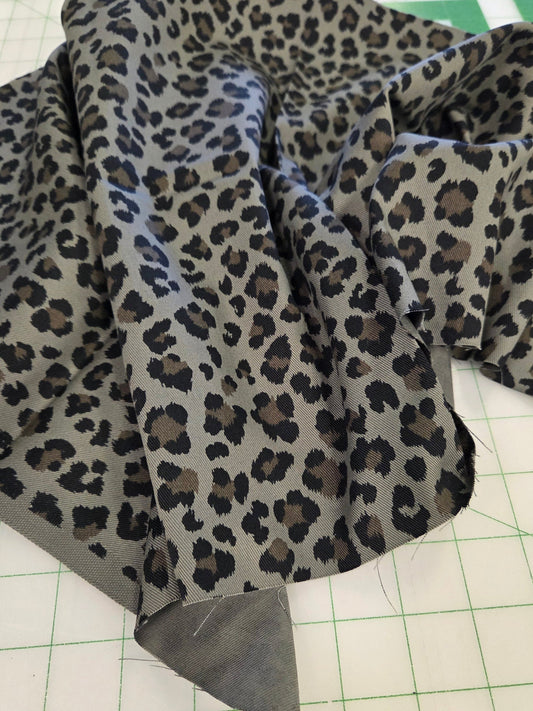 A close-up of leopard-print fabric draped on a white cutting mat with green grid lines. The fabric has a gray base with black and dark brown leopard spots, and a solid gray fabric piece is visible at the folded edge.