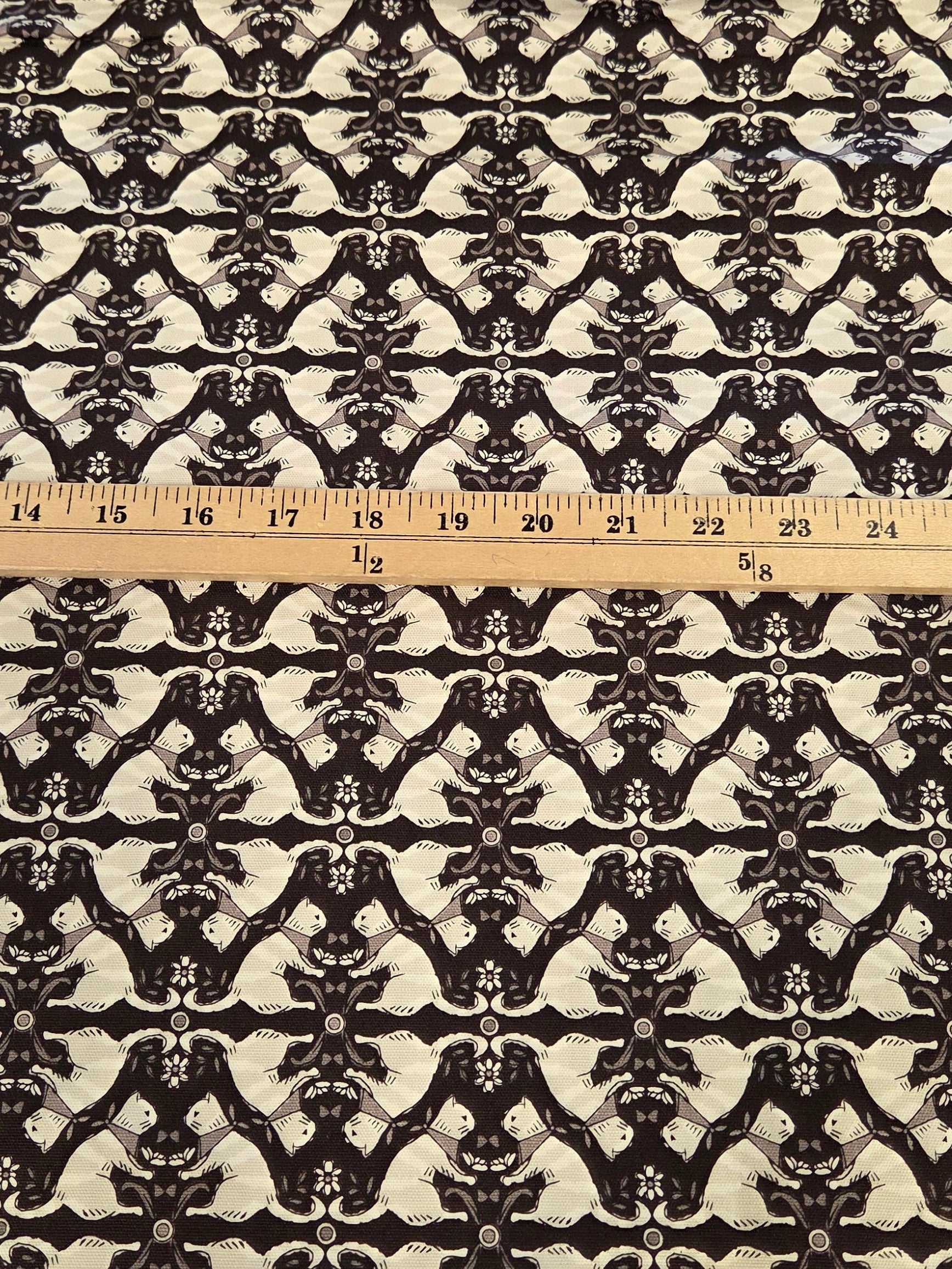 A rectangular piece of fabric with a black and white repeating ornamental damask pattern, laid flat on a green gridded cutting mat with a wooden ruler placed vertically down the center for scale.