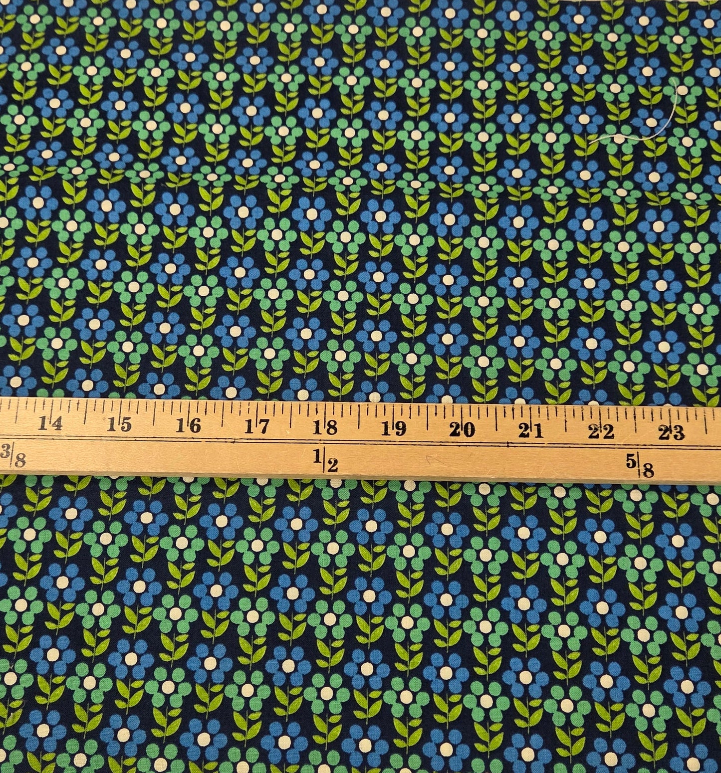 A strip of fabric with a repeating blue, green, and white floral pattern, shown vertically next to a wooden ruler on a green grid cutting mat.