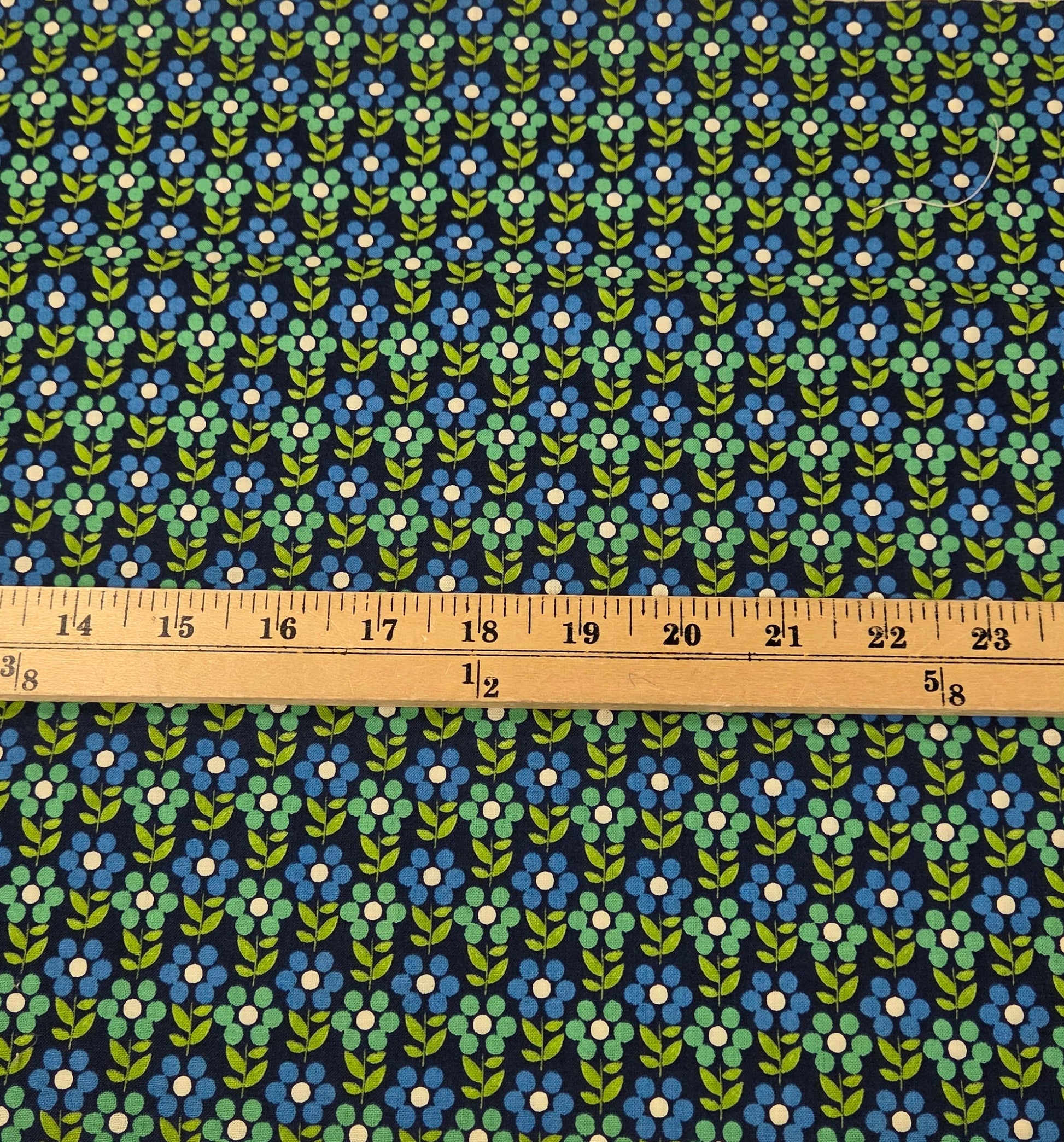 A strip of fabric with a repeating blue, green, and white floral pattern, shown vertically next to a wooden ruler on a green grid cutting mat.