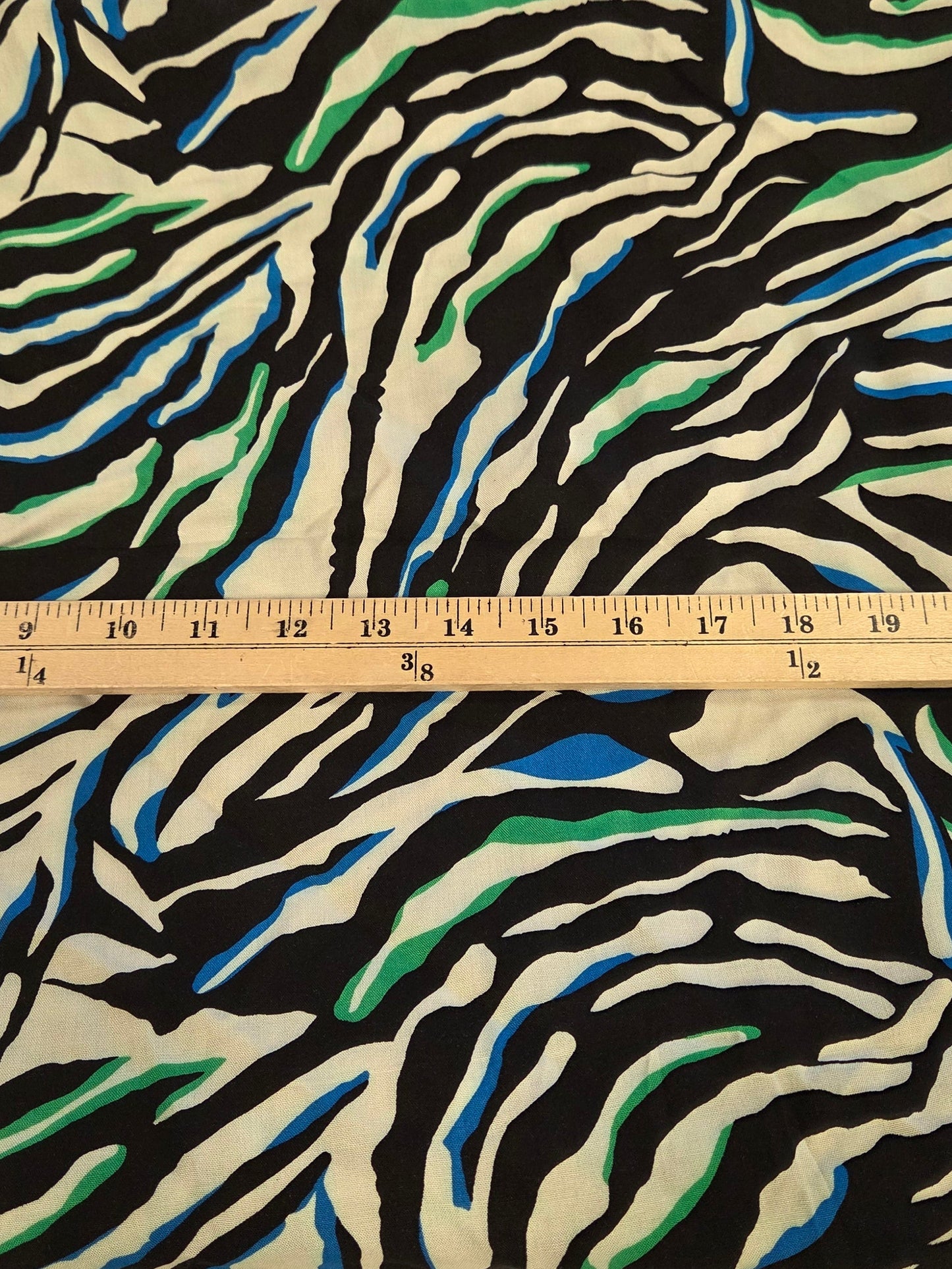 A folded piece of fabric with a zebra-stripe pattern in black, white, blue, and green, shown next to a wooden ruler on a green grid cutting mat.