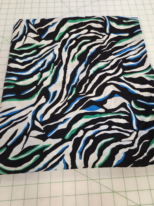 A square fabric sample with a zebra-like abstract pattern in black, white, blue, and green, laid flat on a white grid cutting mat.