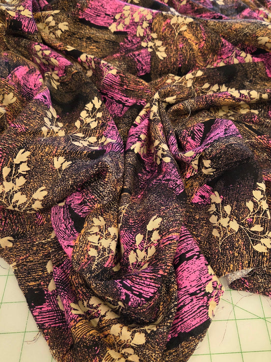 A draped piece of batik fabric with a floral and leaf pattern in pink, purple, brown, and beige, laid out on a green grid cutting mat.