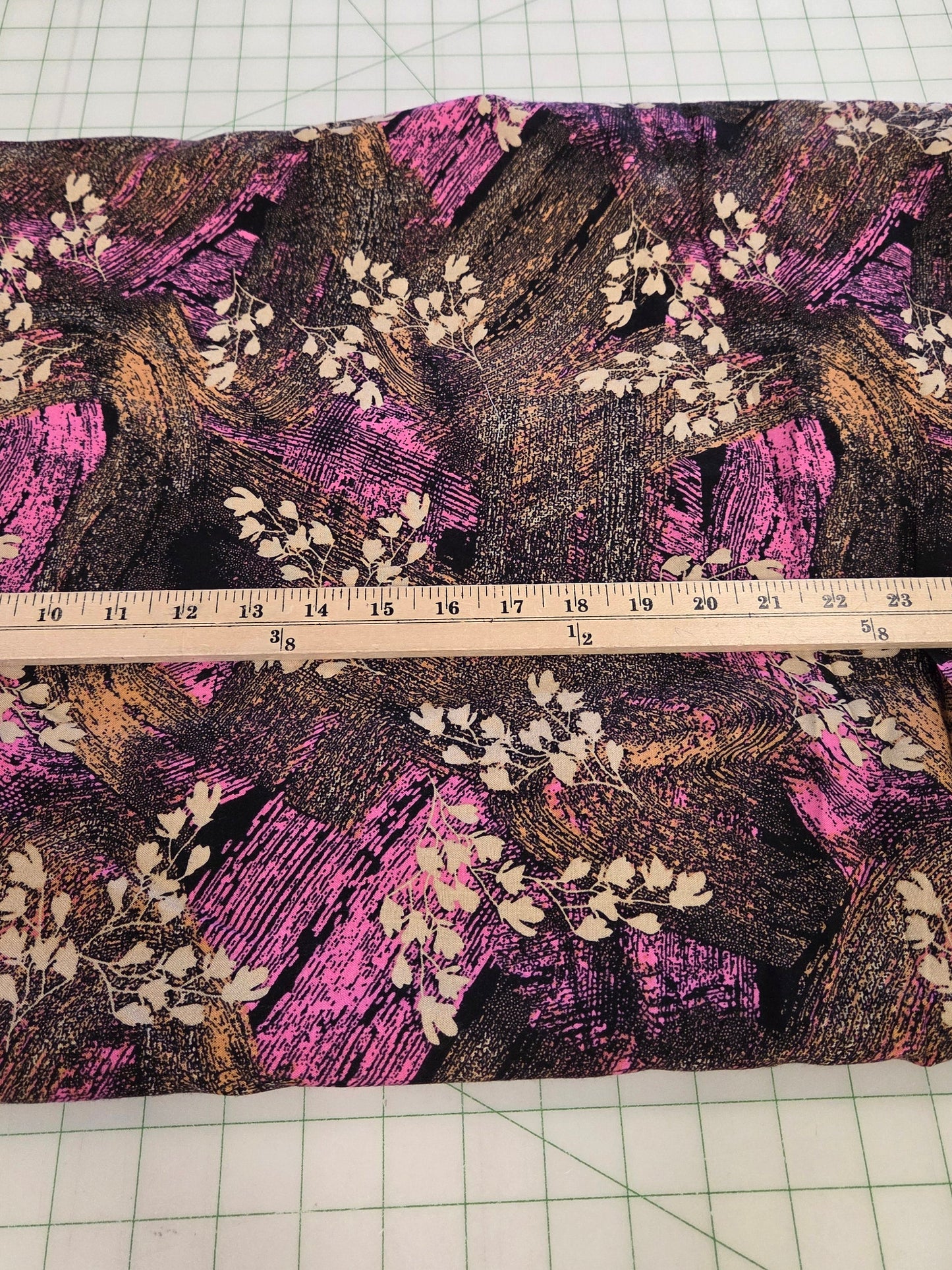 A rectangular piece of fabric with a dark brown and black textured background, decorated with pink brushstroke patterns and white leafy branches, laid flat on a green grid cutting mat with a wooden ruler across the middle.