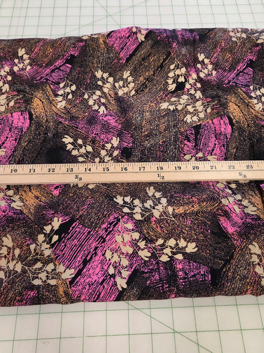 A rectangular piece of fabric with a dark brown and black textured background, decorated with pink brushstroke patterns and white leafy branches, laid flat on a green grid cutting mat with a wooden ruler across the middle.
