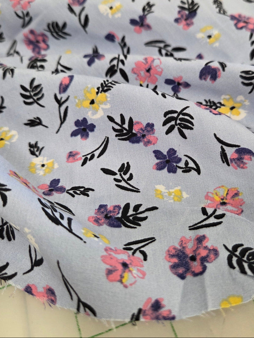 Light blue fabric with a delicate floral and leaf print in pink, yellow, purple, and black, draped over a green-grid cutting mat.