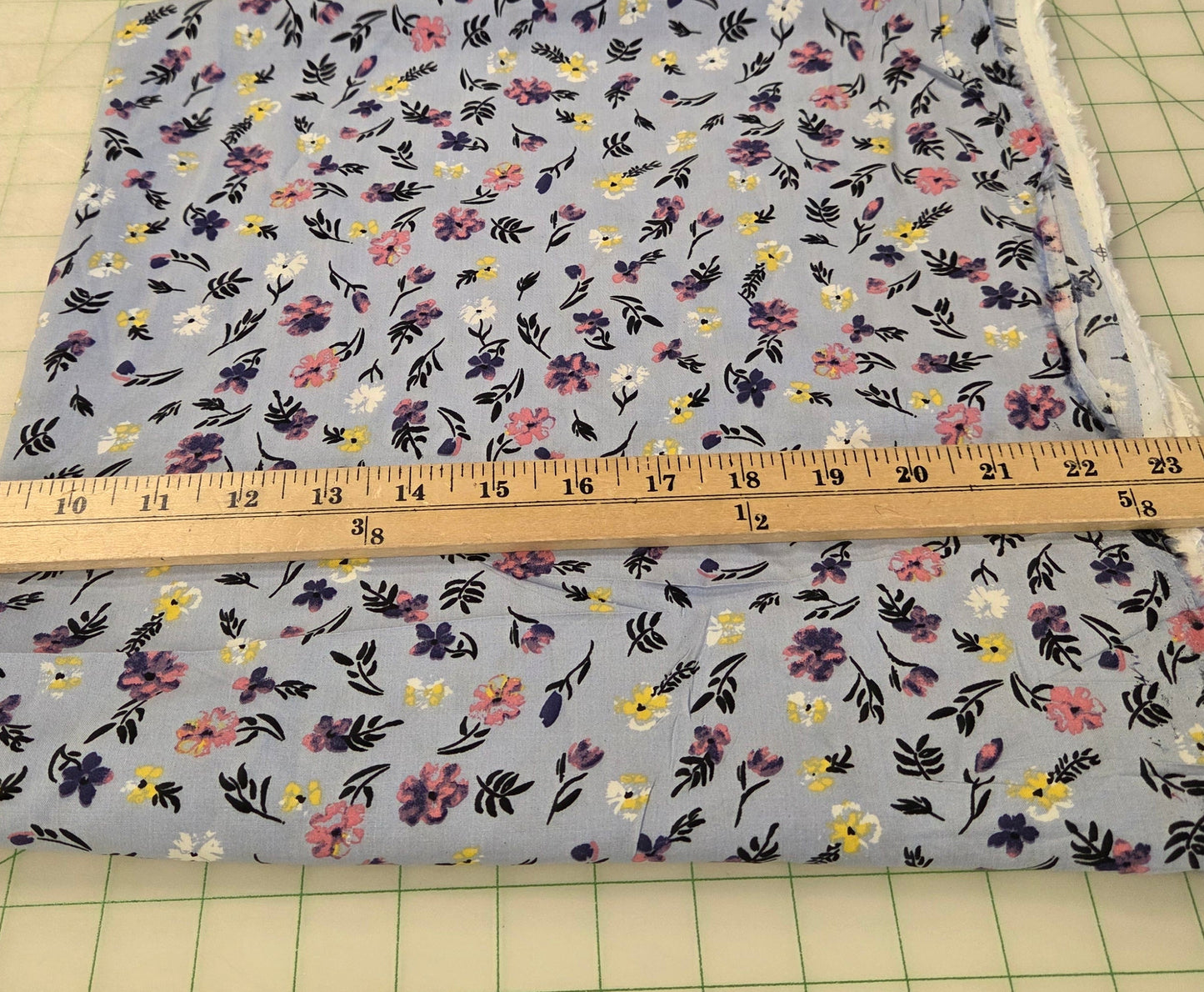 A square piece of light blue floral fabric with a small-scale multicolor print, shown next to a wooden yardstick on a green grid cutting mat.
