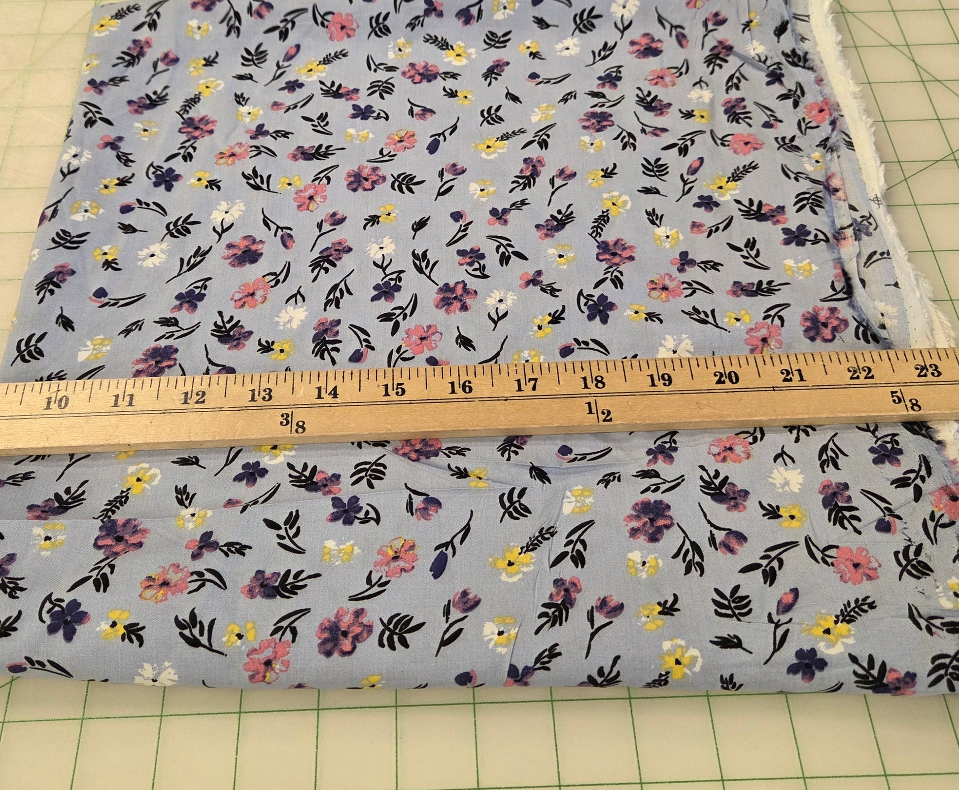 A square piece of light blue floral fabric with a small-scale multicolor print, shown next to a wooden yardstick on a green grid cutting mat.