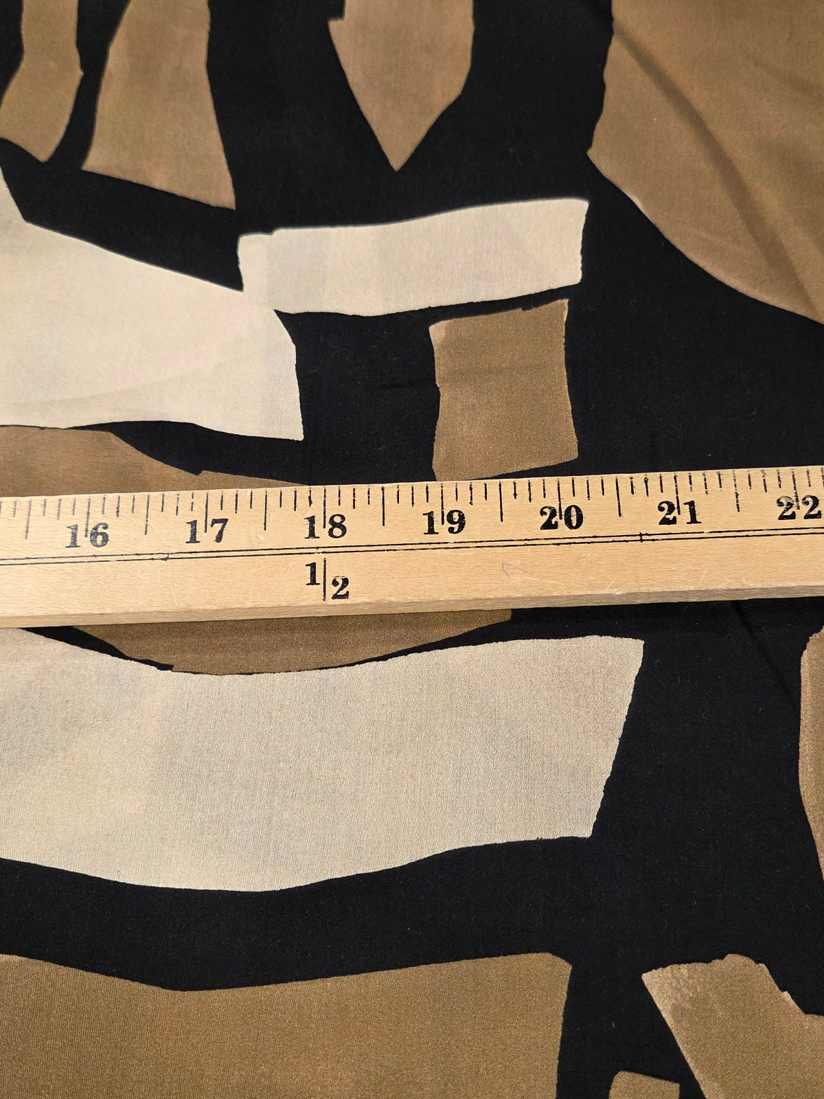 A close-up of a fabric with an abstract geometric pattern in black, beige, and white, shown next to a wooden ruler for scale.