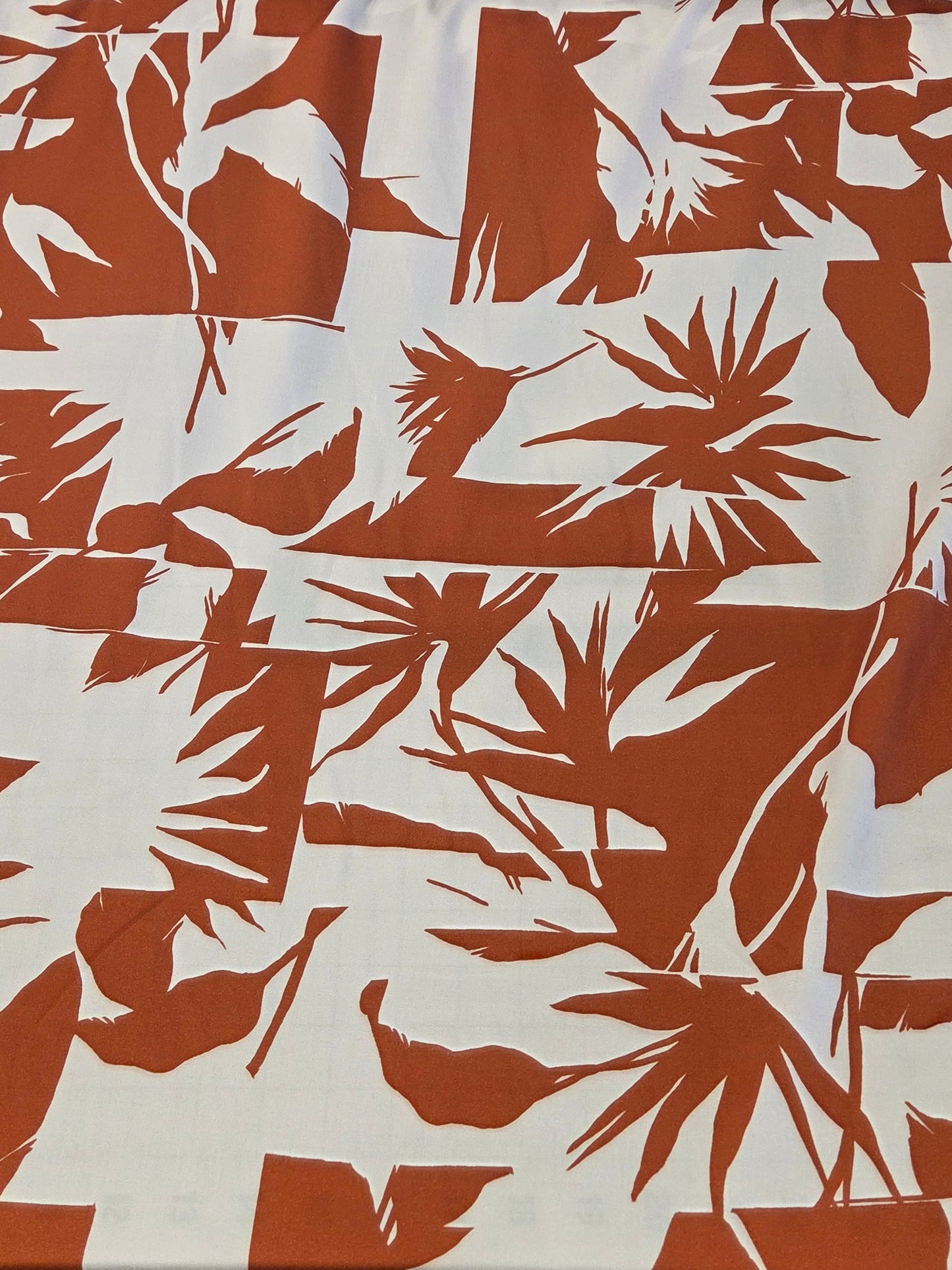 A close-up of a fabric or wallpaper sample featuring a bold, repeating tropical leaf pattern in terracotta orange on a white background.