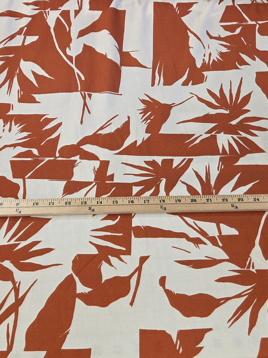 A close-up of patterned fabric with large rust-brown tropical leaf silhouettes on an off-white background, shown with a wooden ruler running vertically for scale.