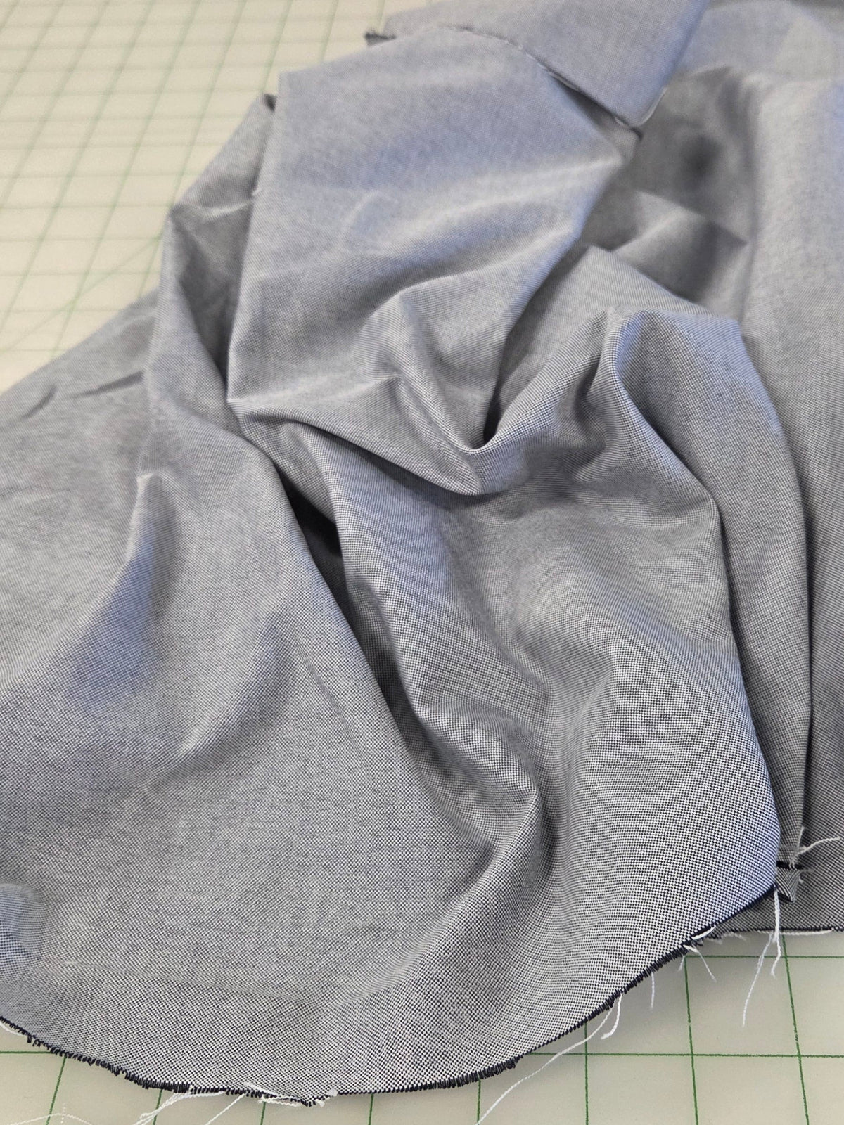 Light blue fabric draped over a green grid cutting mat in a sewing or tailoring workspace, with sewing tools and other fabric visible in the background.