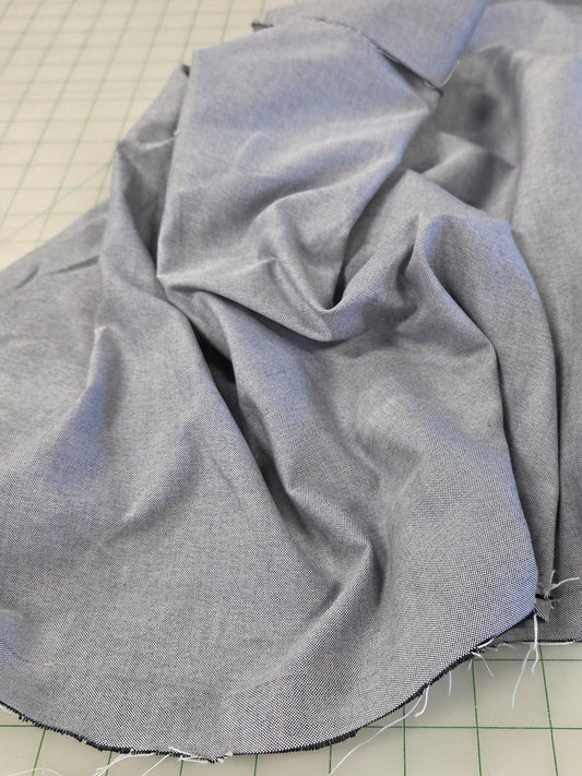 Light blue fabric draped over a green grid cutting mat in a sewing or tailoring workspace, with sewing tools and other fabric visible in the background.