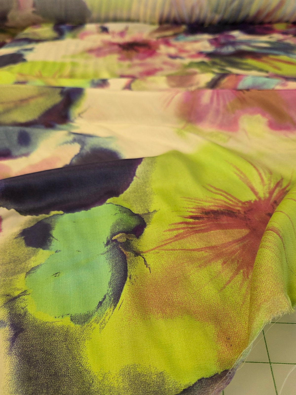 A close-up of a colorful fabric with a watercolor floral pattern in lime green, pink, purple, and blue, draped next to a white cutting mat with grid lines.
