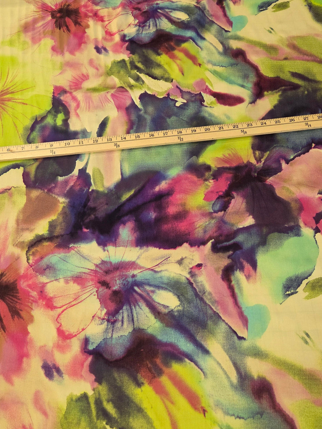 A close-up of a colorful floral fabric with a watercolor-style print in pink, green, blue, purple, and yellow on a light cream base, with a measuring tape laid across it for scale.