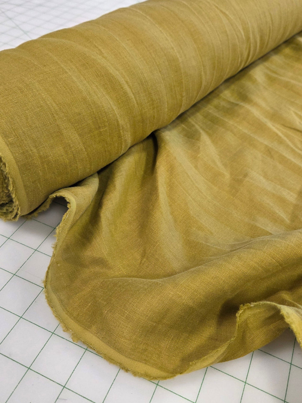 A roll of textured olive green fabric partially unrolled on a white gridded cutting mat in a sewing or fabric workshop.