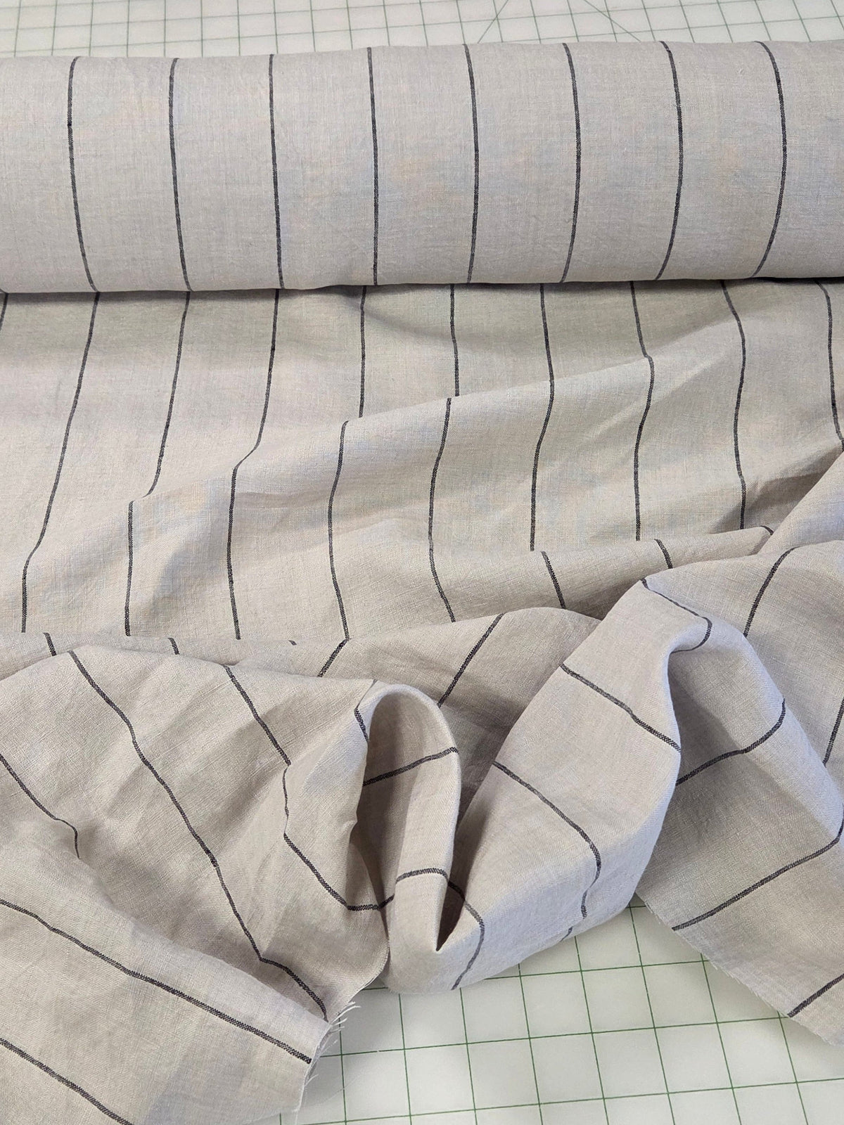 A roll of light gray fabric with thin black horizontal stripes laid out on a white gridded cutting mat, with a small tan fabric sample and scissors visible at the left edge.
