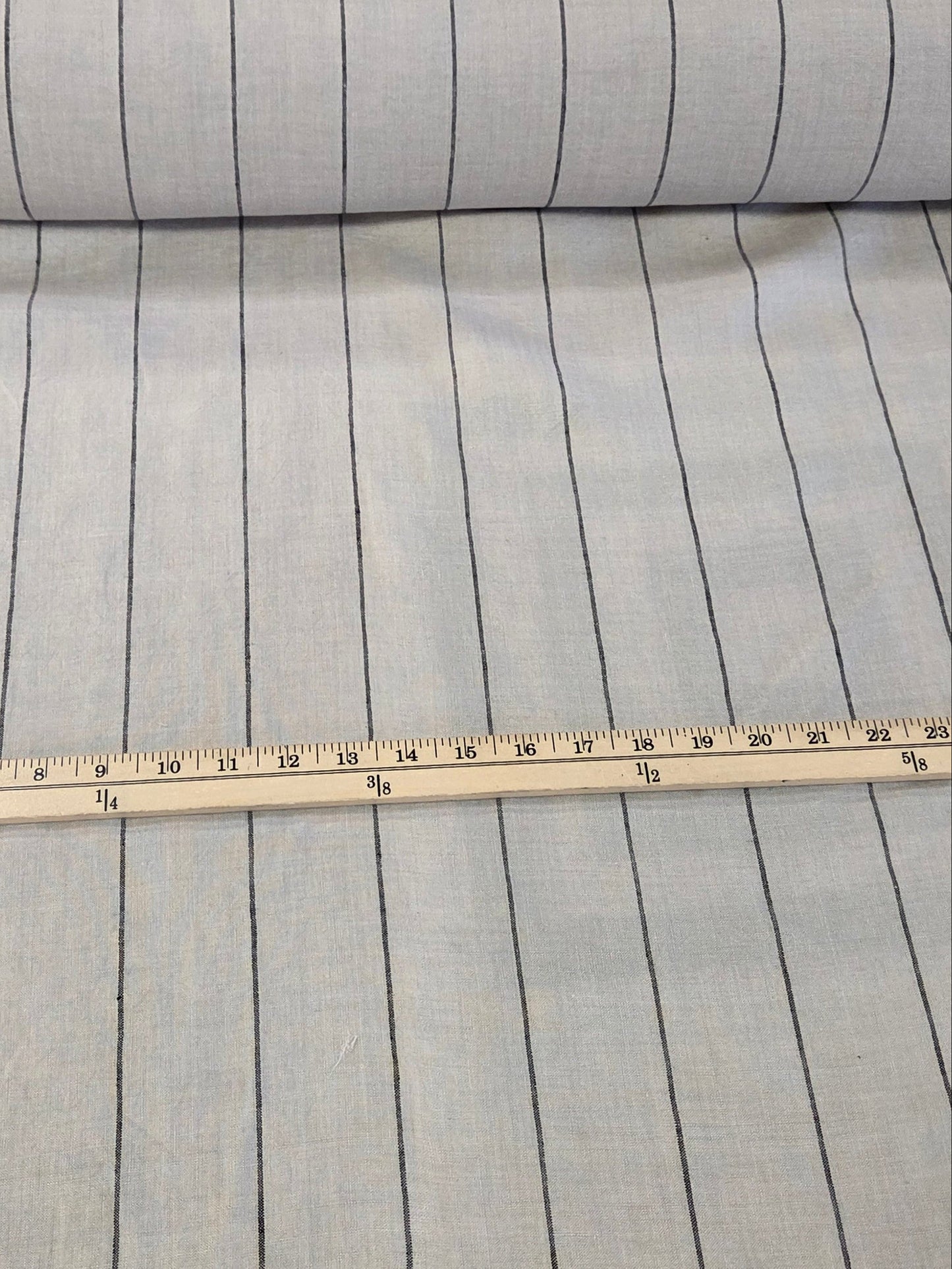 A roll of light gray fabric with thin black horizontal stripes, shown next to a wooden ruler for scale on a cutting mat.