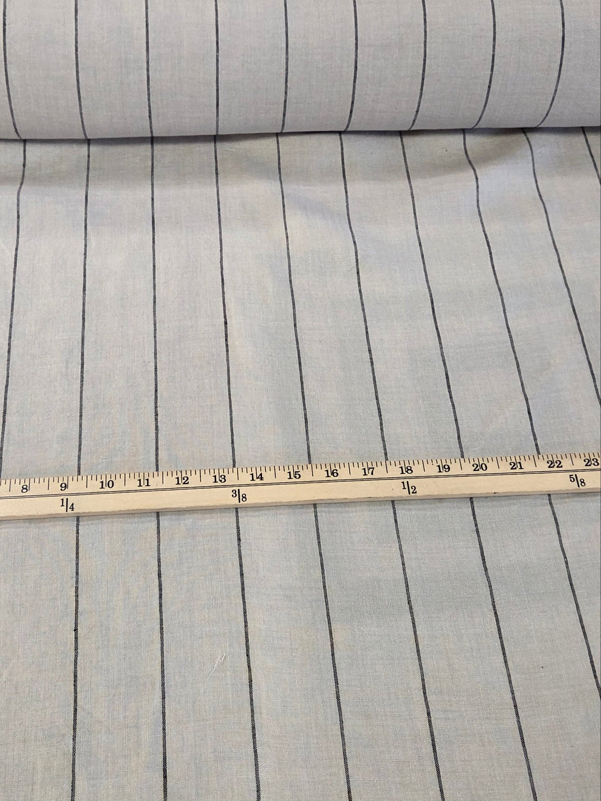 A roll of light gray fabric with thin black horizontal stripes, shown next to a wooden ruler for scale on a cutting mat.