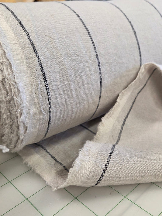 A close-up of a rolled piece of light beige fabric with thin black horizontal stripes, showing its woven texture and raw frayed edges, resting on a white cutting mat with a green grid.