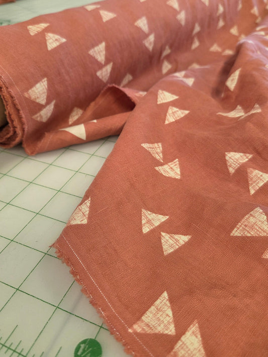 A close-up of terracotta-colored fabric with a repeating white geometric triangle pattern, draped over a white gridded cutting mat.