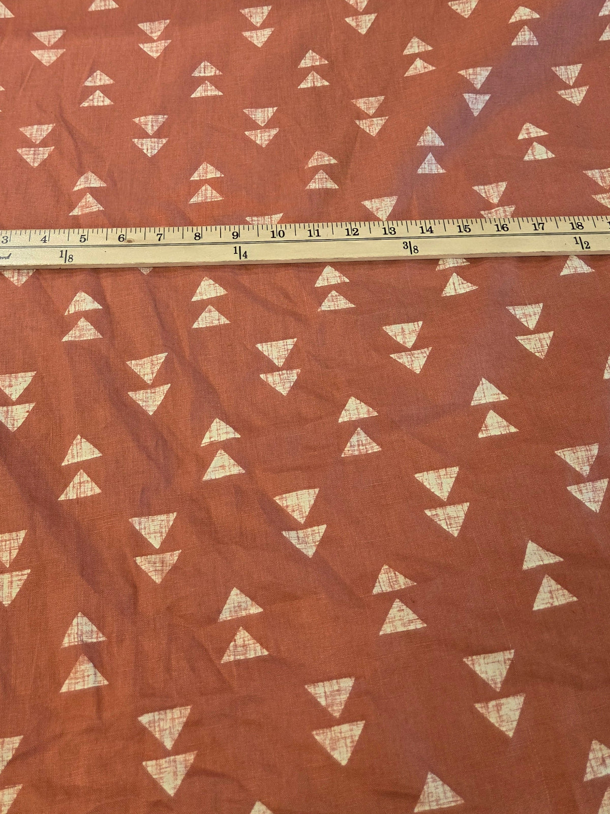 Terracotta orange fabric with a repeating white triangle pattern, laid out on a cutting mat with a wooden ruler measuring its width.
