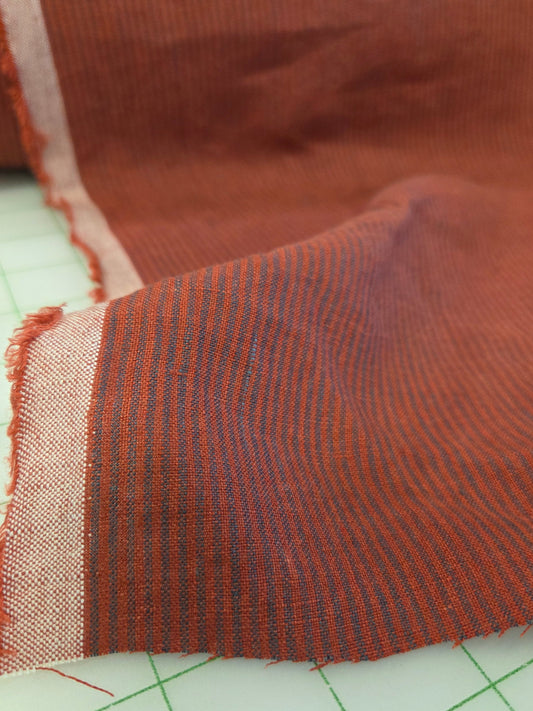 A close-up of a woven fabric swatch in a rust-brown color with fine dark stripes and a lighter beige border, laid on a green grid cutting mat.