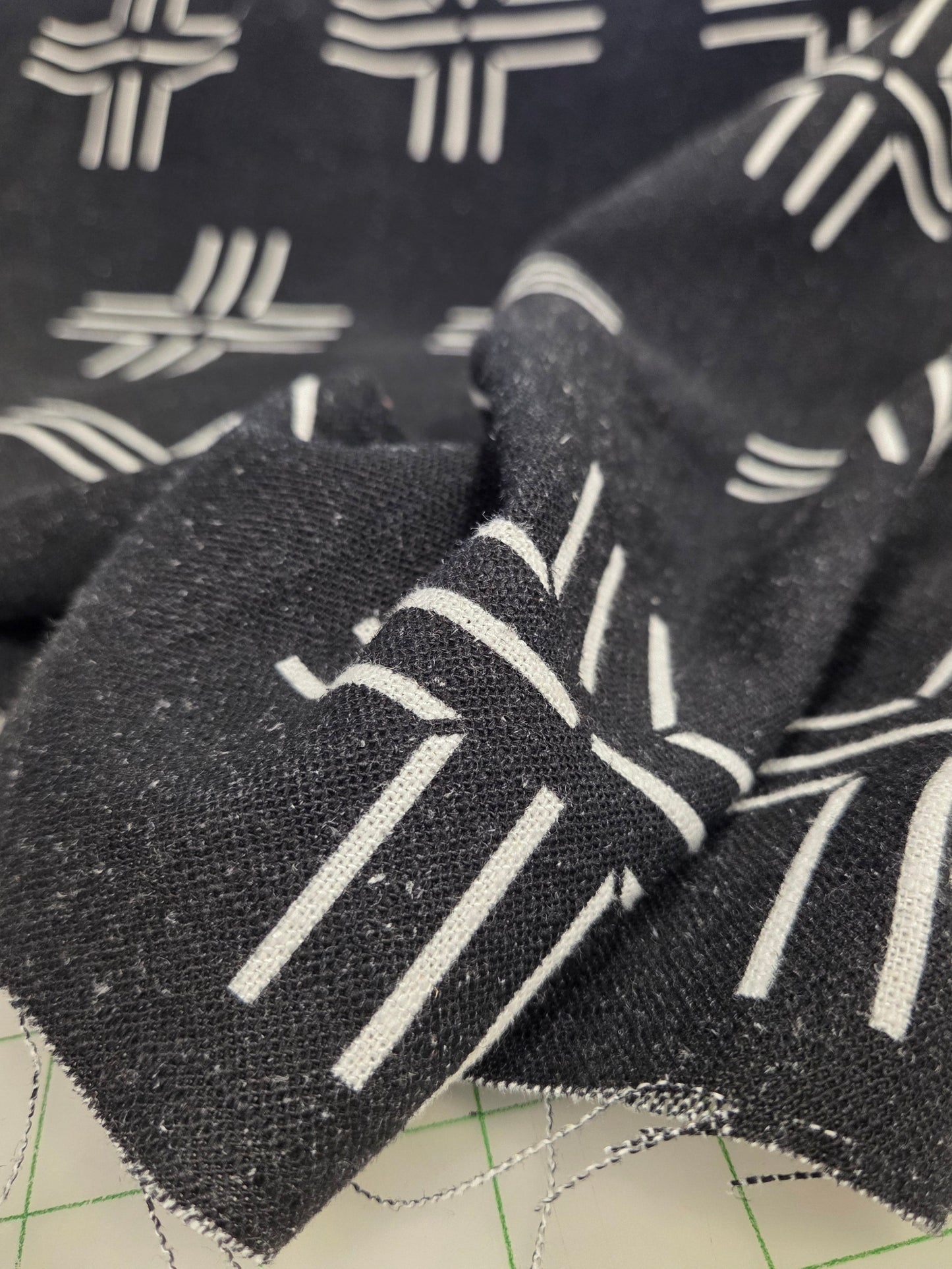 A close-up of a black woven fabric with a white geometric pattern, draped over a beige cutting mat with green grid lines.