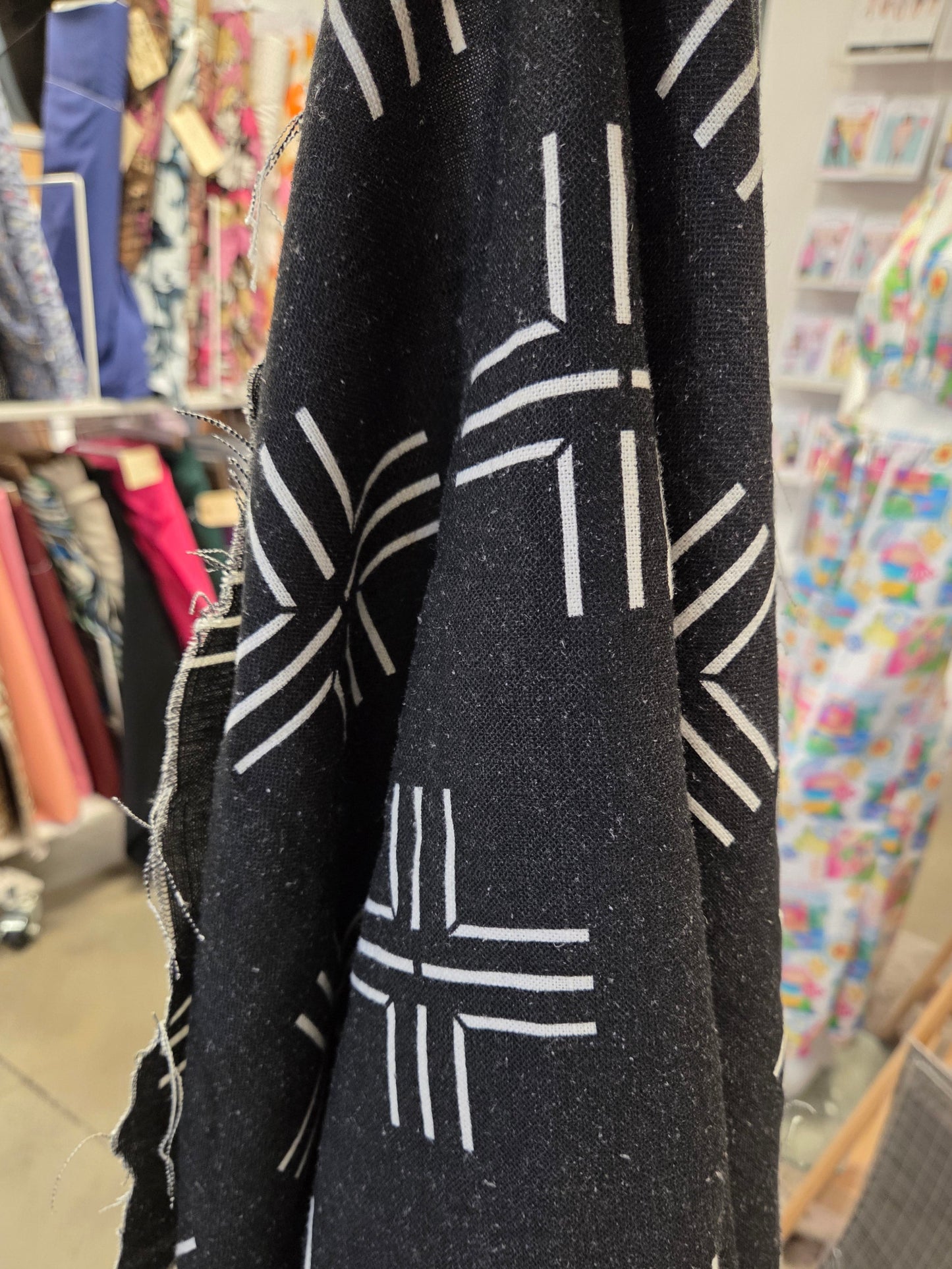 A close-up of a black fabric with a white geometric dash pattern, draped in the foreground with a blurred shop or textile store background.