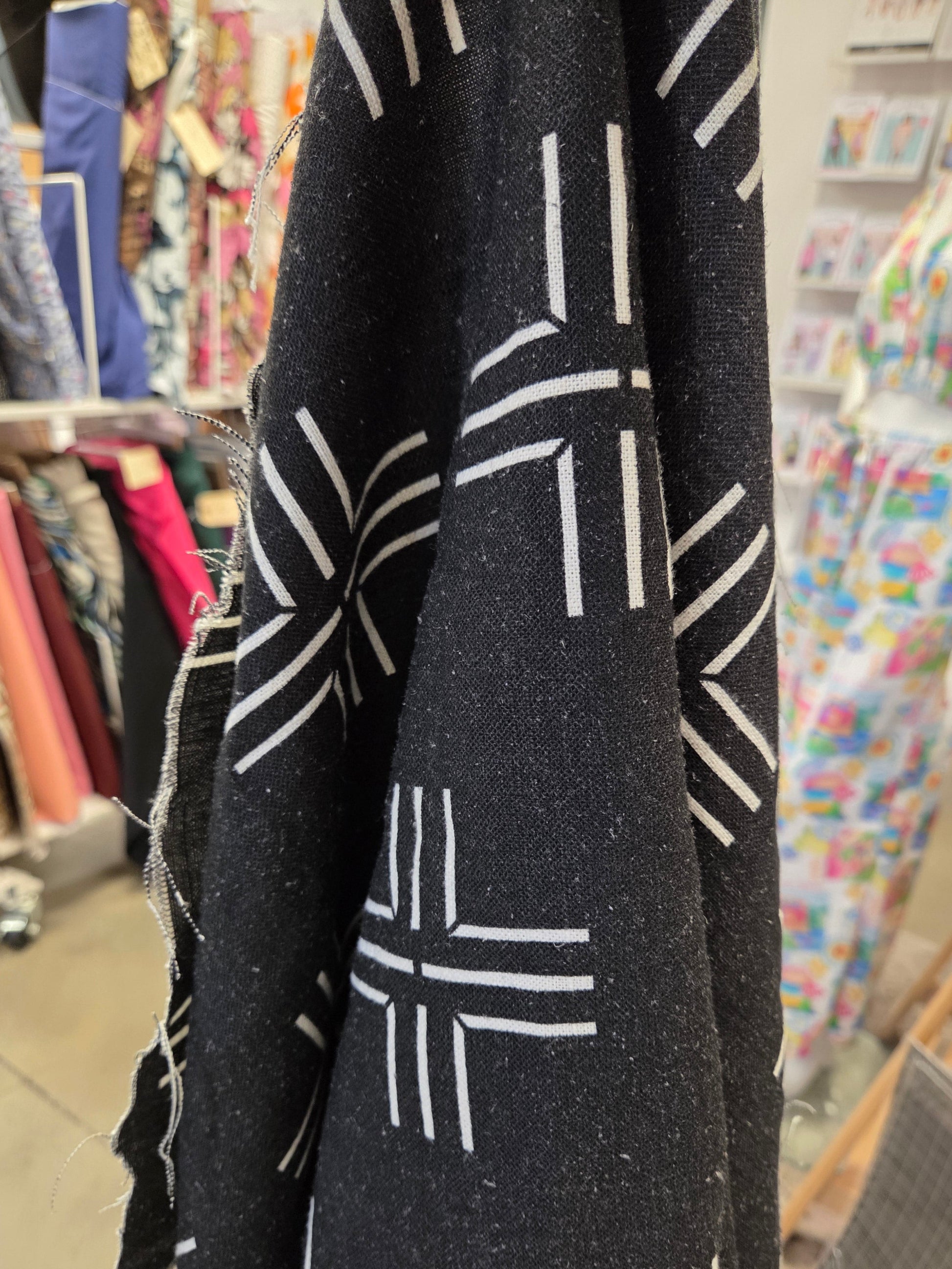 A close-up of a black fabric with a white geometric dash pattern, draped in the foreground with a blurred shop or textile store background.