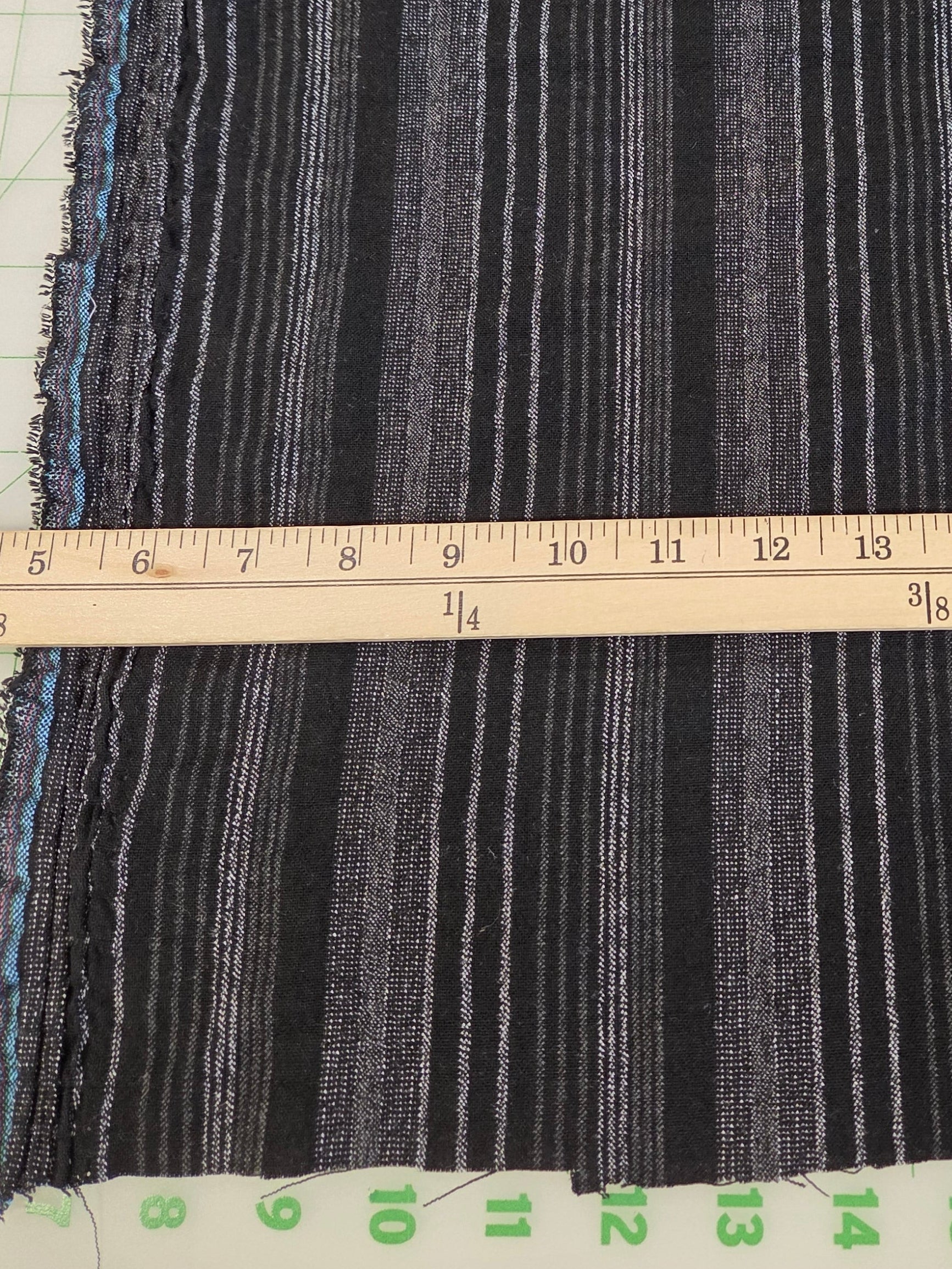 A close-up of striped fabric with alternating black, gray, and white horizontal lines, shown with a wooden ruler and green measurement grid for scale.