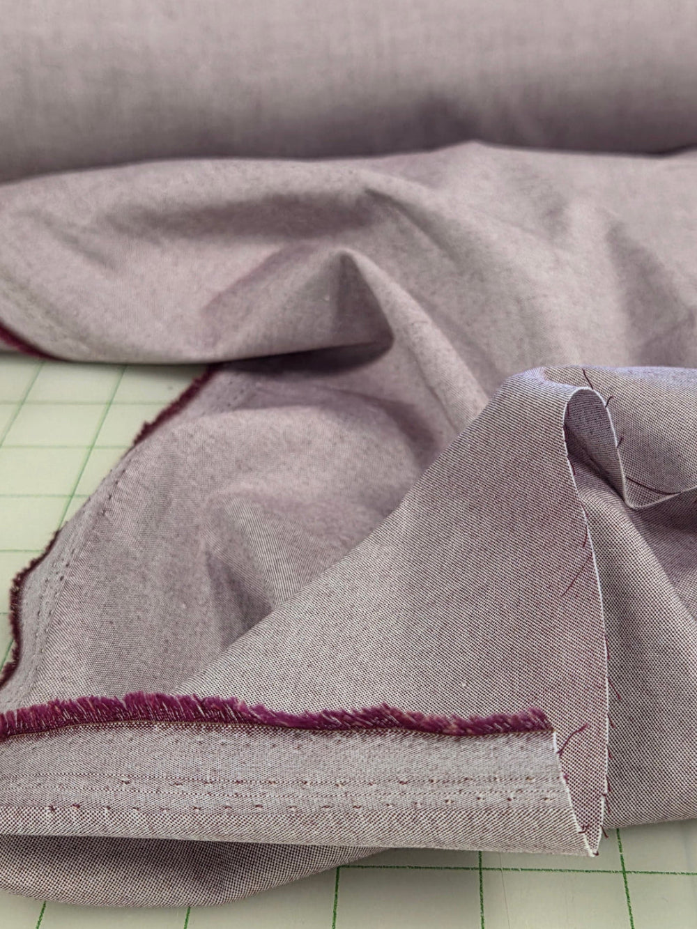 A close-up of a light gray fabric garment or textile with a folded cuff showing a dark burgundy stitched edge, set against a white gridded background.