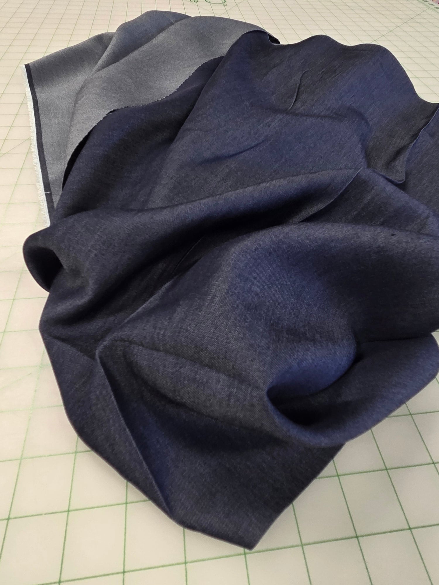 A dark navy blue fabric garment, likely a skirt or draped textile, laid out on a white gridded cutting mat with shelves of colorful fabrics visible in the background.
