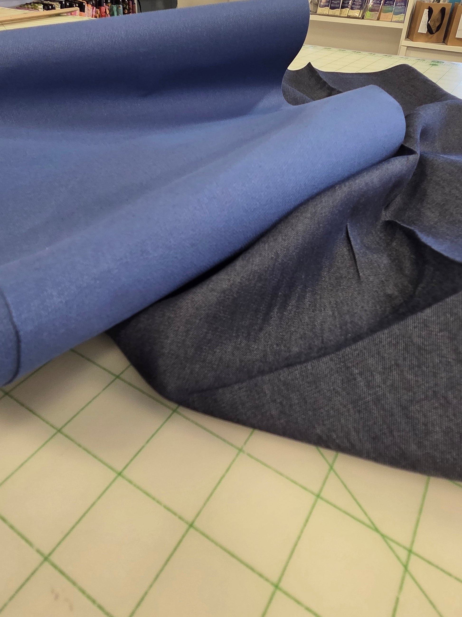 A roll of medium blue fabric partially unrolled onto a light-colored cutting mat inside a sewing or fabric store, with shelves and sewing machines visible in the background.