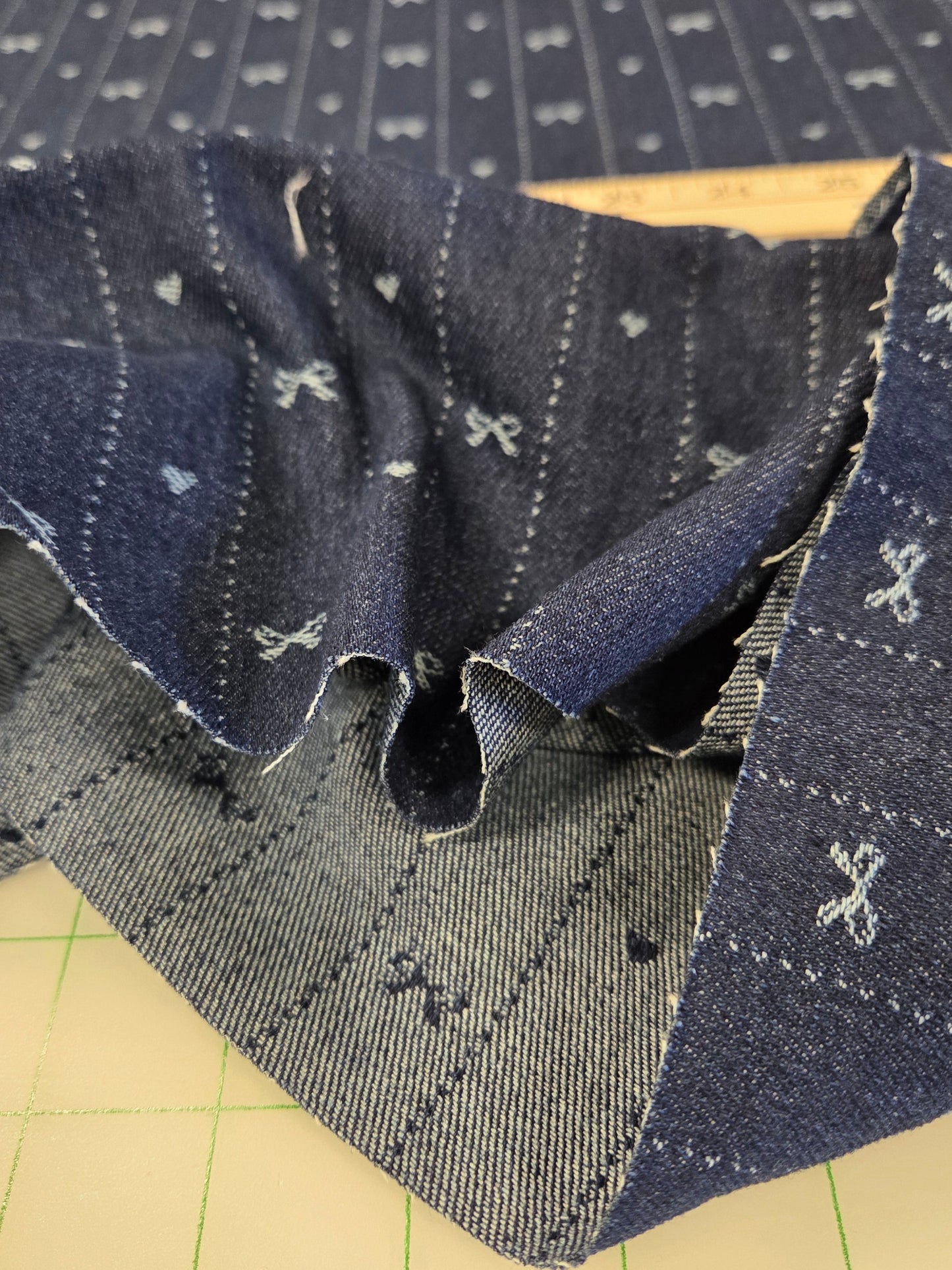 A close-up of a dark blue denim fabric with small white embroidered motifs, showing both the front patterned side and the reverse textured side, laid on a beige cutting mat with green grid lines.