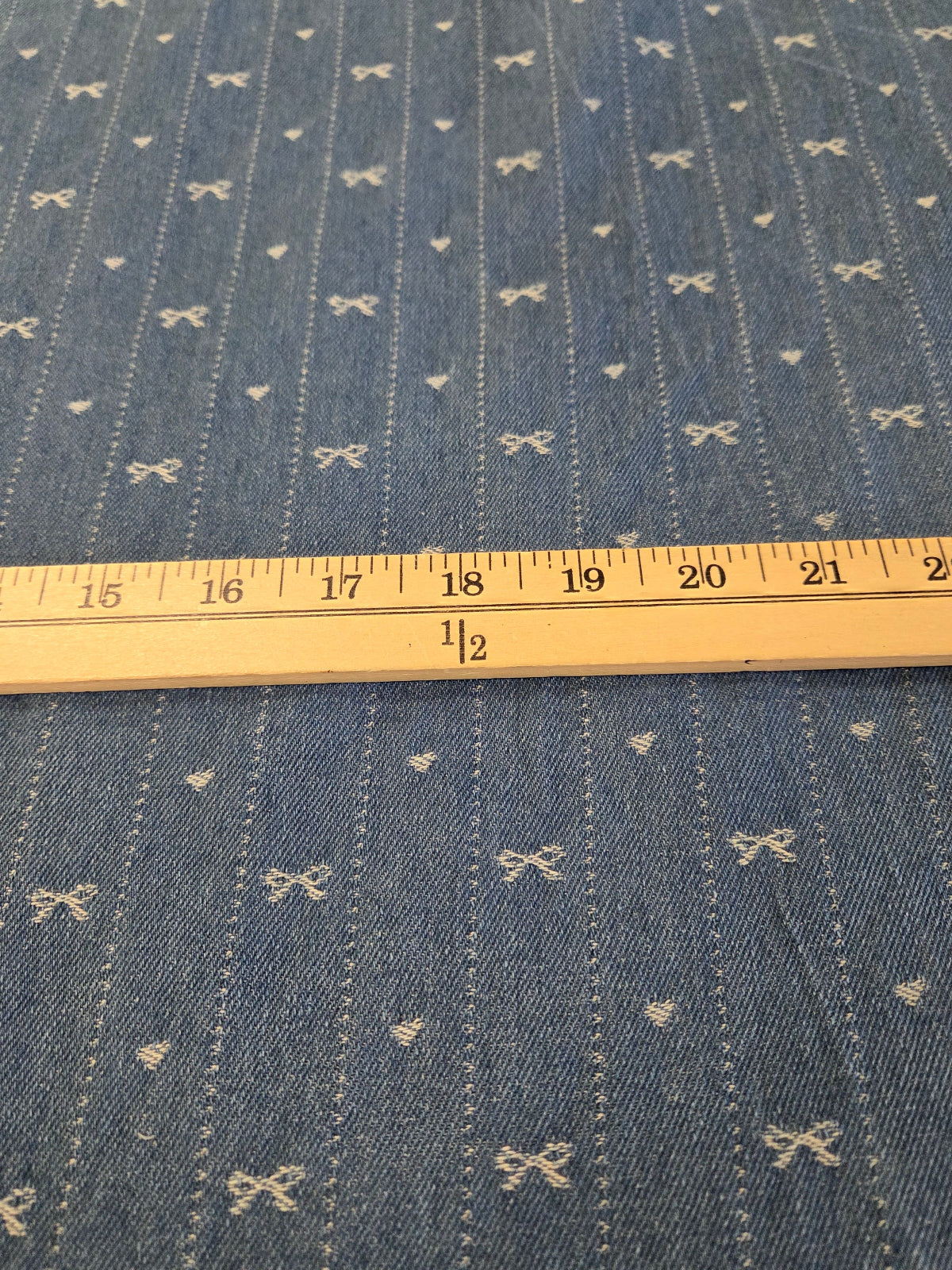 Blue denim fabric with a repeating pattern of small white bow and heart motifs, shown with a wooden ruler for scale.