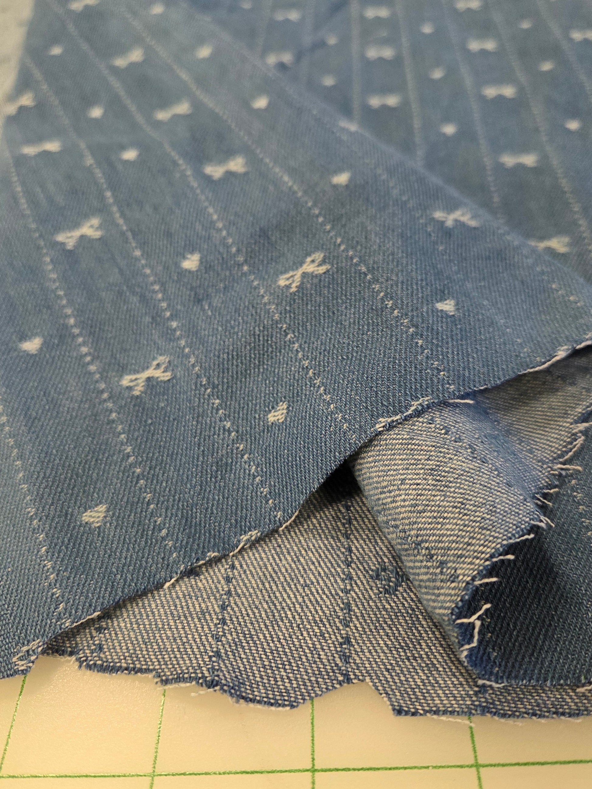 A close-up of folded blue denim fabric with a small white embroidered pattern, resting on a green-lined cutting mat.