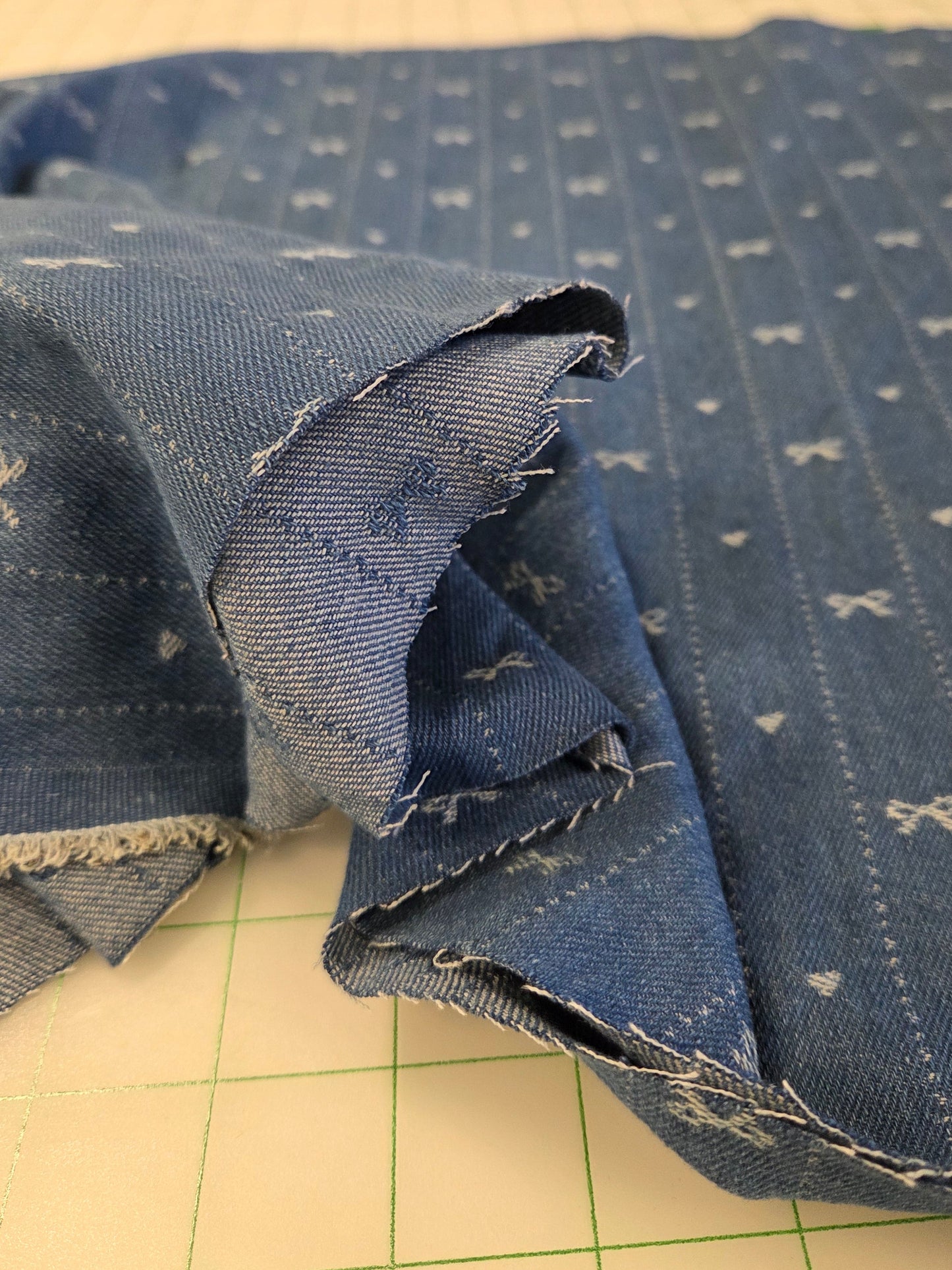 A close-up of blue denim fabric with a white bow and dot pattern, folded over on a beige cutting mat.