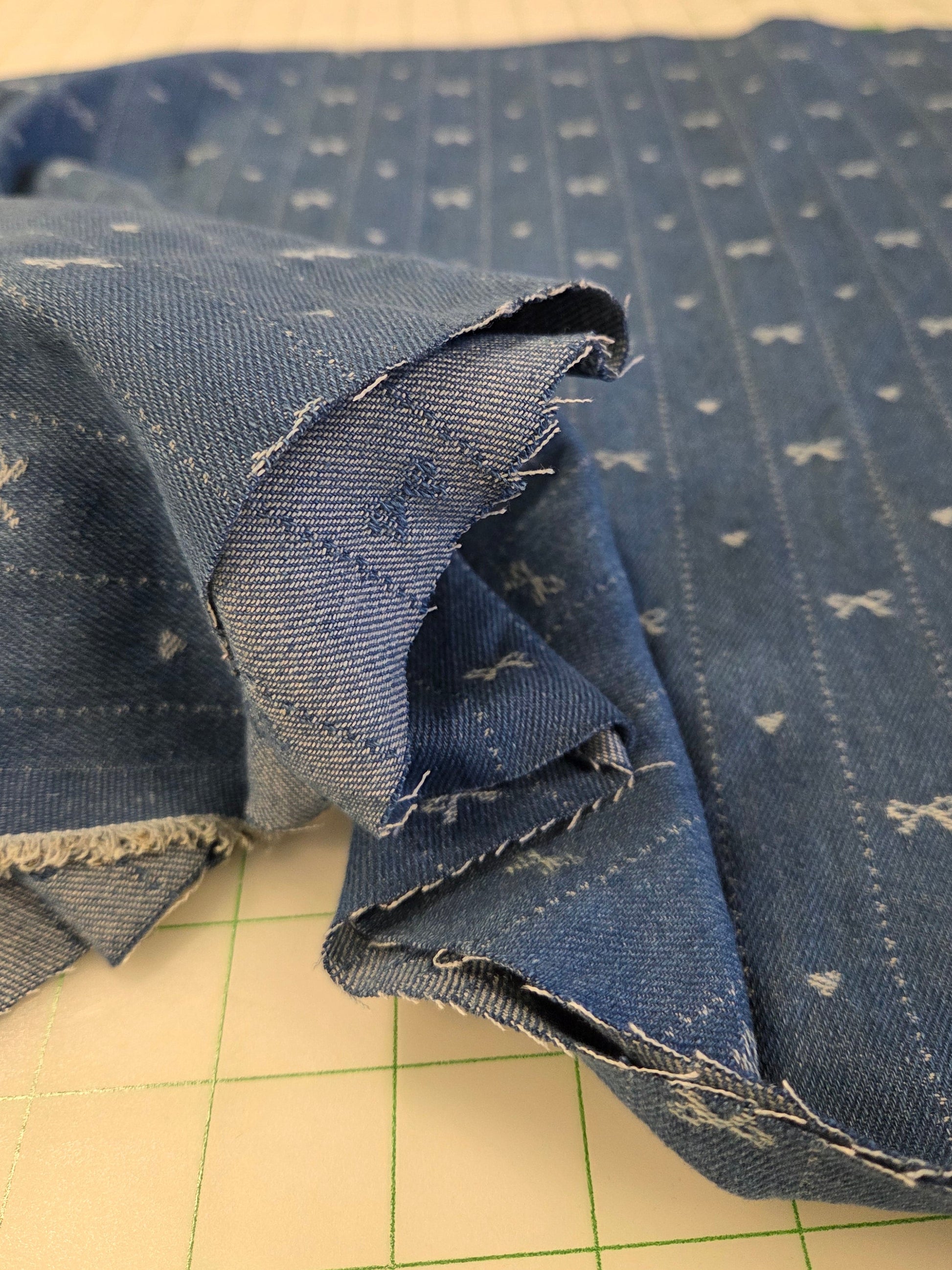 A close-up of blue denim fabric with a white bow and dot pattern, folded over on a beige cutting mat.
