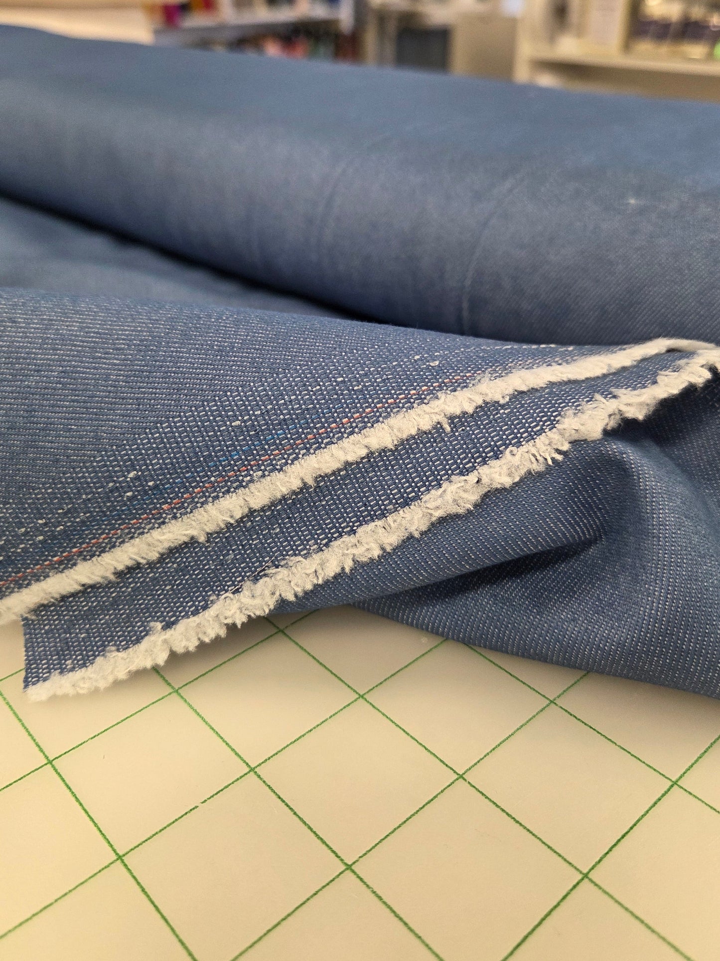 A close-up of a piece of blue denim fabric with a raw, frayed white edge, draped over a cutting mat in a sewing or fabric shop.
