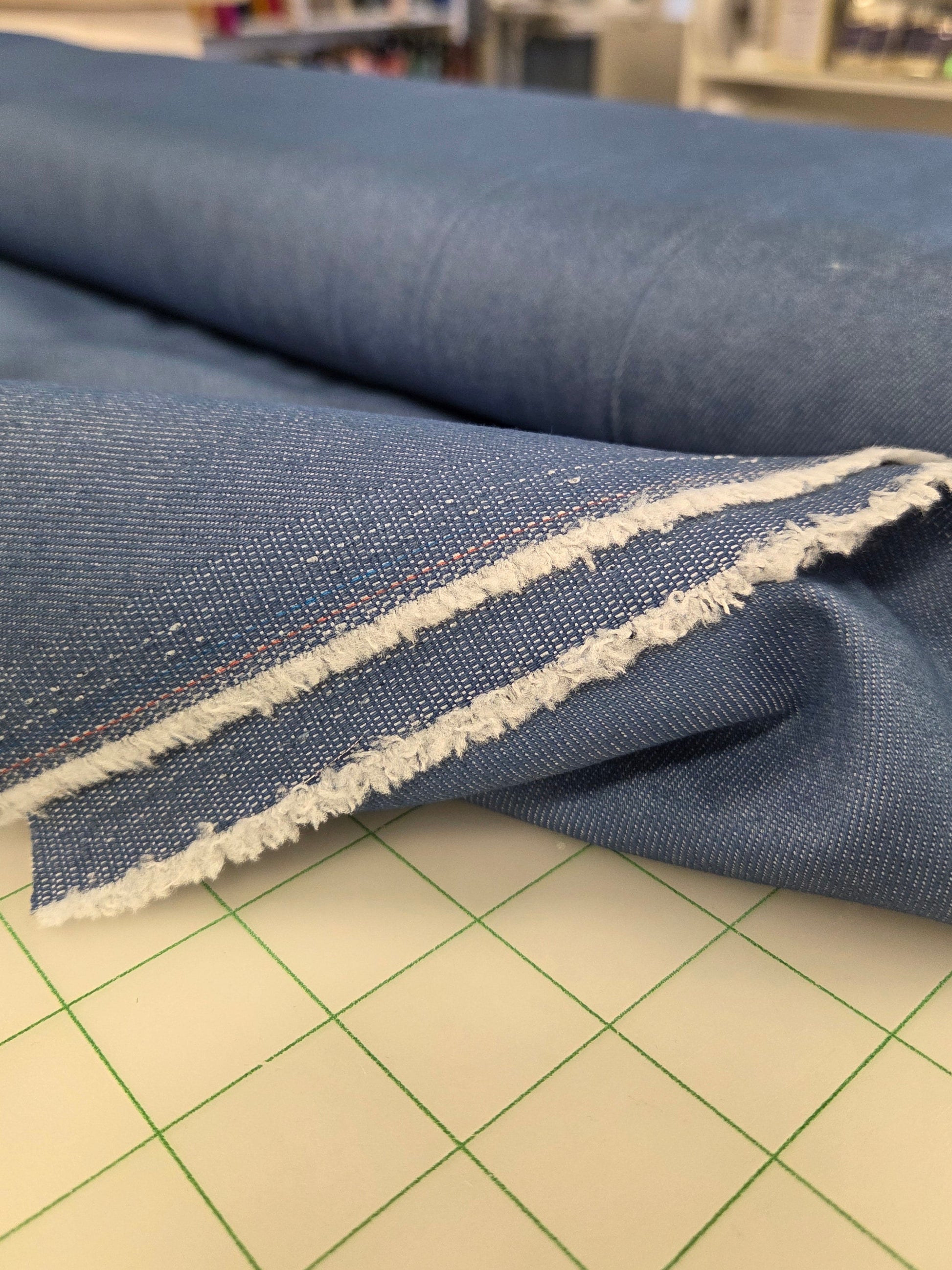 A close-up of a piece of blue denim fabric with a raw, frayed white edge, draped over a cutting mat in a sewing or fabric shop.