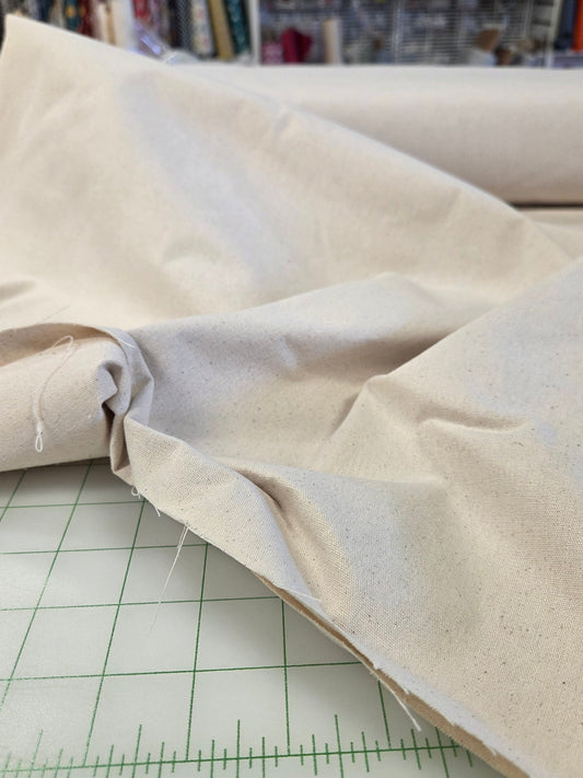 A close-up of a light beige fabric roll draped over a white cutting mat with green measurement markings, with shelves of sewing supplies visible in the background.
