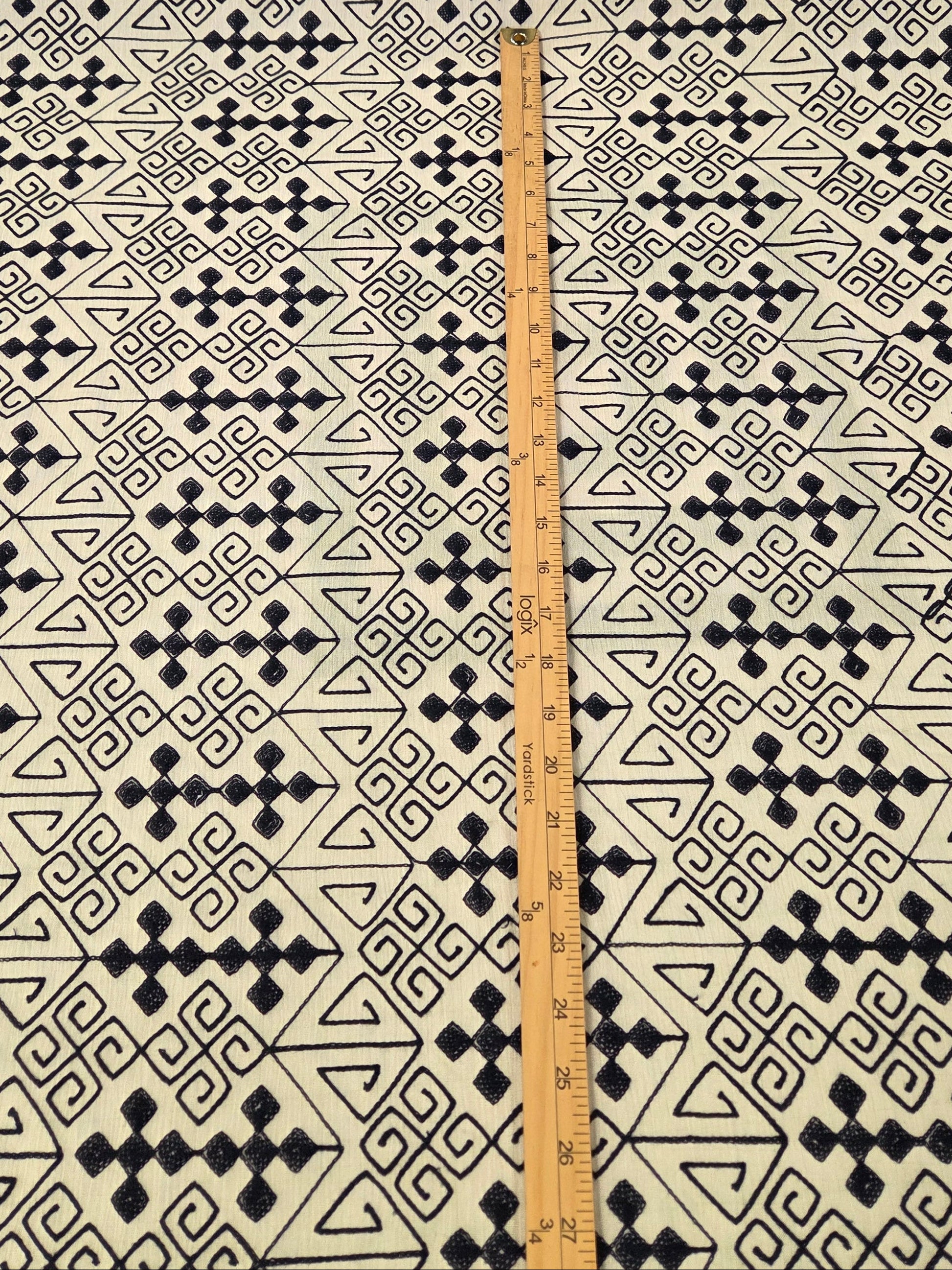 A close-up of a blue geometric patterned fabric, with a wooden ruler placed vertically in the center to show the scale of the pattern.