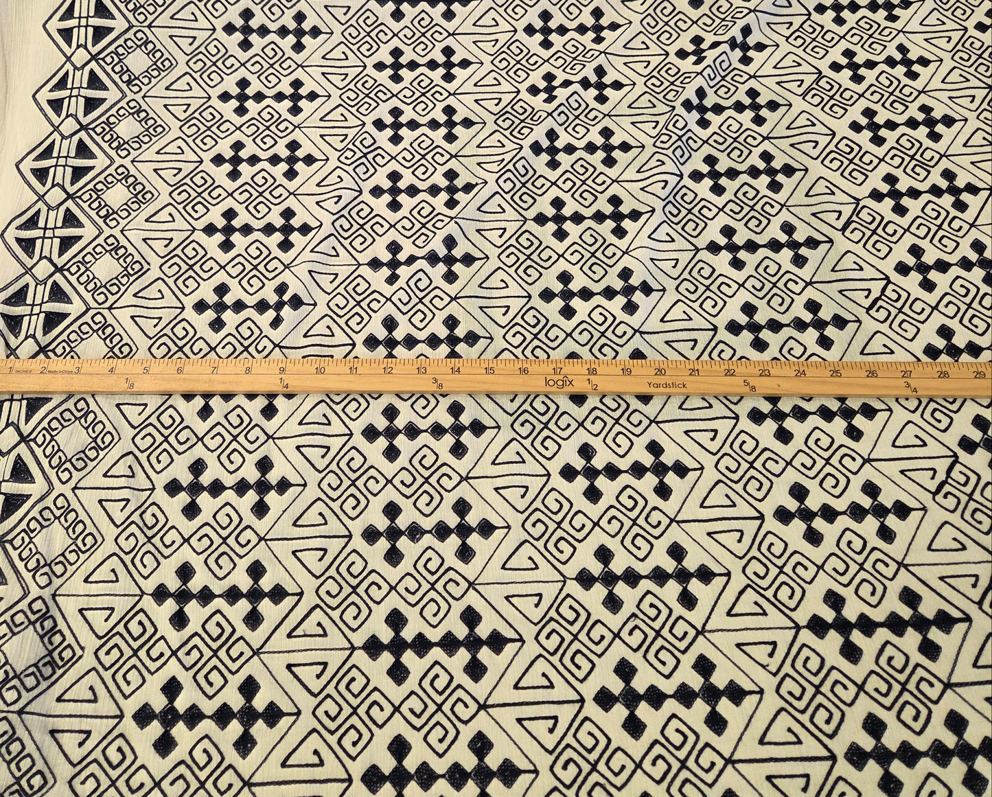 A large roll of fabric with a black geometric tribal pattern on a white base, laid out horizontally with a wooden ruler placed across the center to show scale.