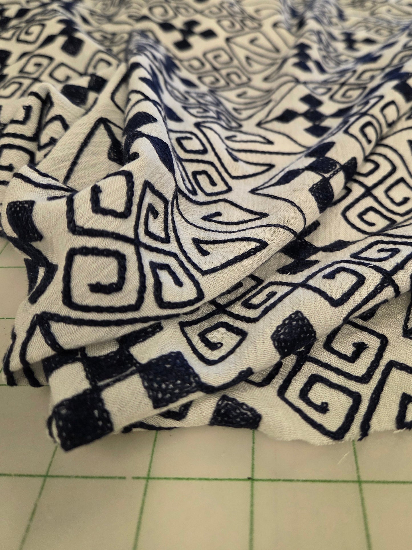 A close-up of draped fabric with a black geometric hand-drawn pattern on a white background, resting on a green-tiled cutting mat.