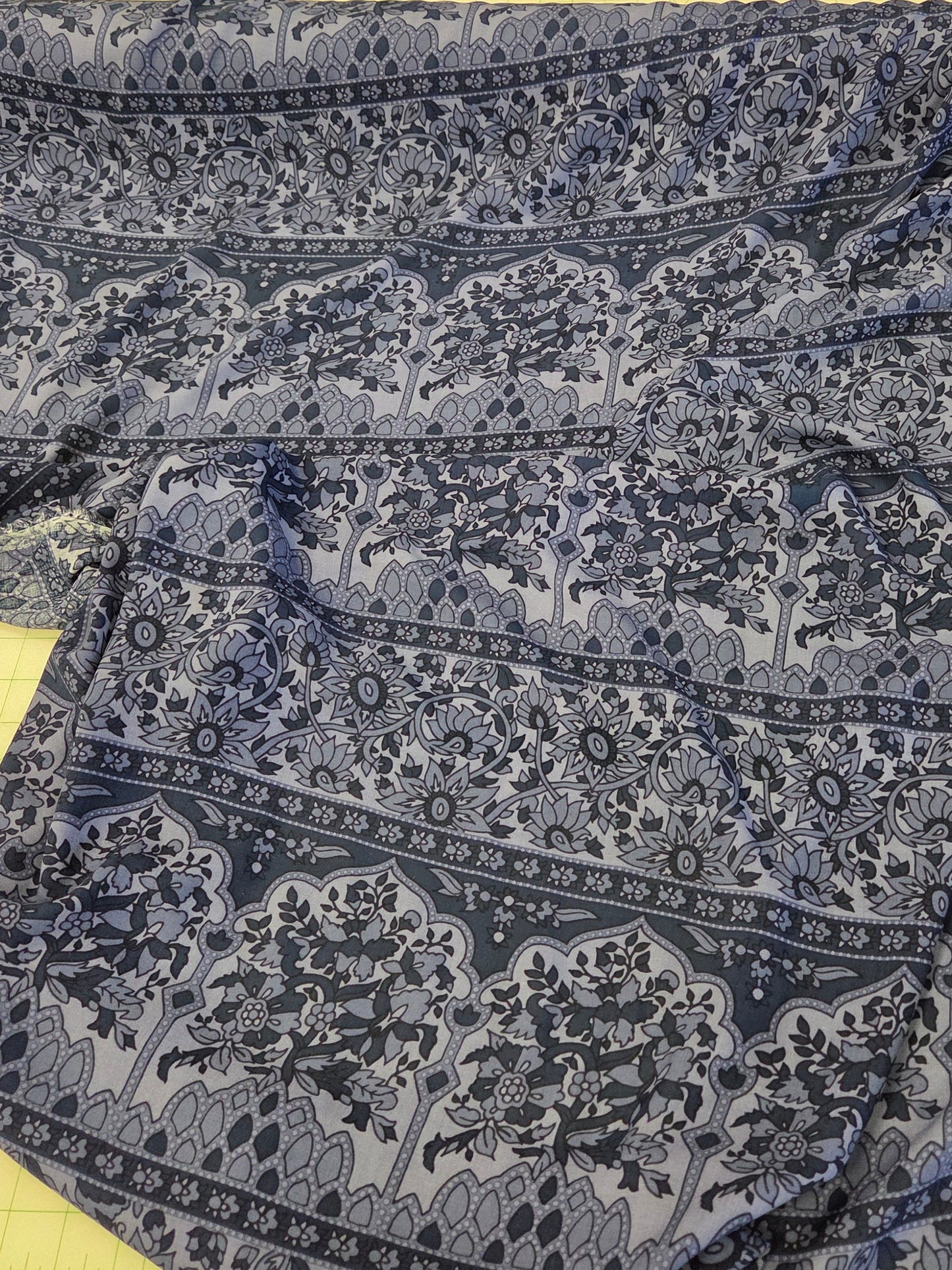 A blue and navy patterned fabric garment laid flat on a cutting mat, with a black fabric roll and measuring tape visible in the background.