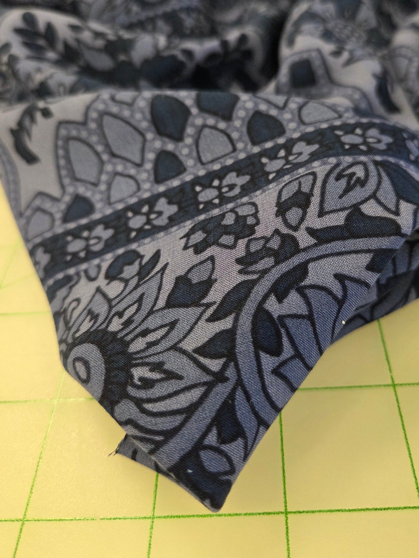 A close-up of a folded piece of fabric with a detailed navy blue and gray floral mandala pattern, resting on a yellow cutting mat with green grid lines.