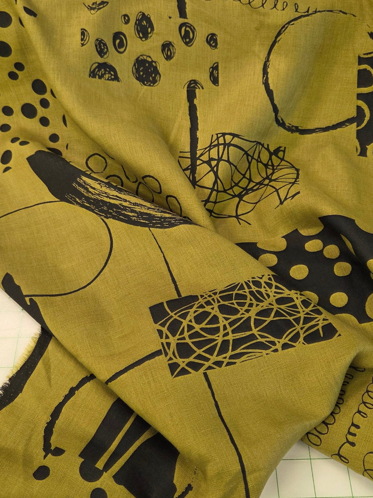 A piece of olive green fabric with a black abstract print, laid out on a white gridded cutting mat.