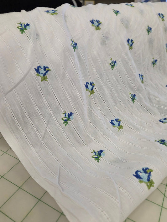 White muslin fabric with a small blue and green floral pattern, draped on a cutting mat in a craft room setting.